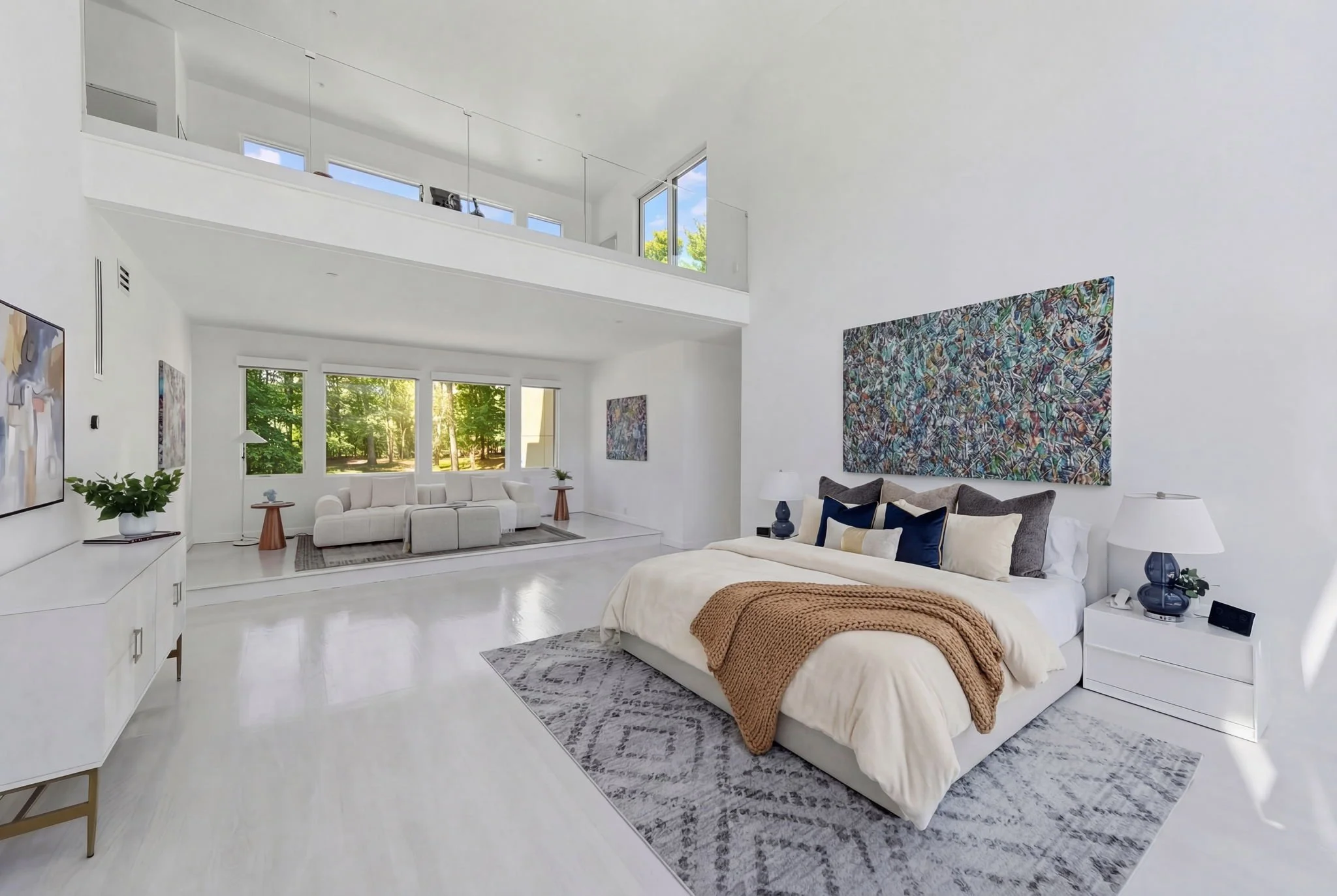 A spacious, modern bedroom with white walls and large windows showing green trees outside. There is a white bed with dark and light blue pillows, a brown knit blanket, and two white nightstands with lamps. An abstract painting hangs above the bed, an