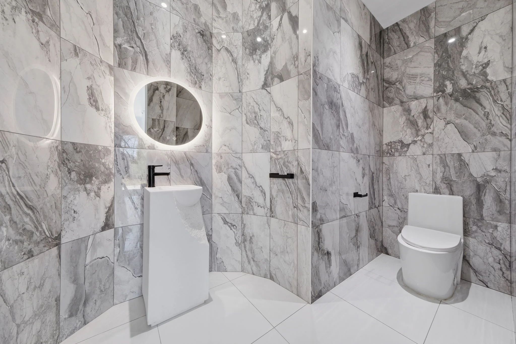 Modern bathroom with marble walls, small white pedestal sink with black faucet, illuminated circular mirror, and a white toilet.