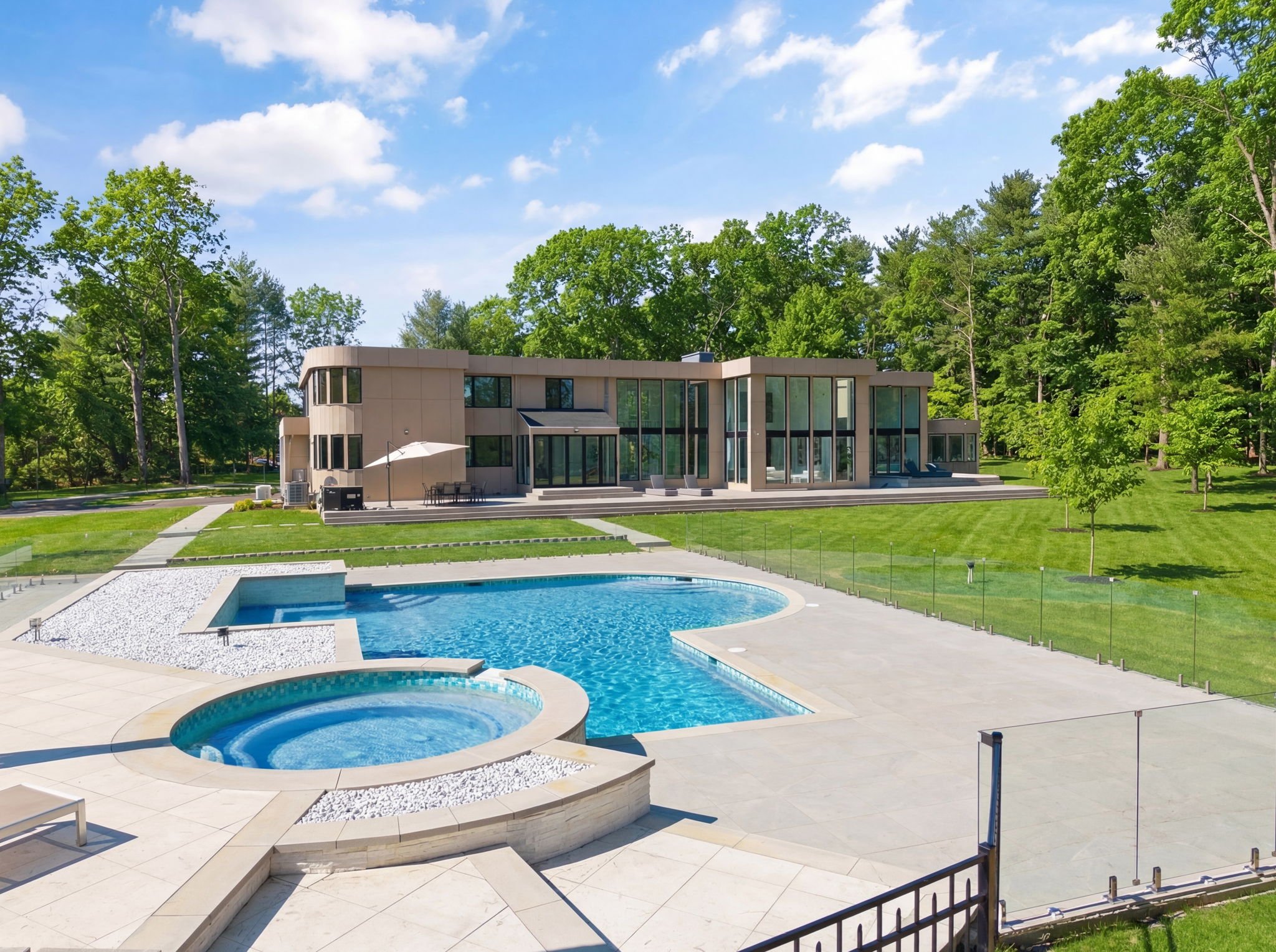 A modern house with large glass windows overlooking a backyard with a swimming pool and hot tub, surrounded by a well-maintained lawn and tall trees.
