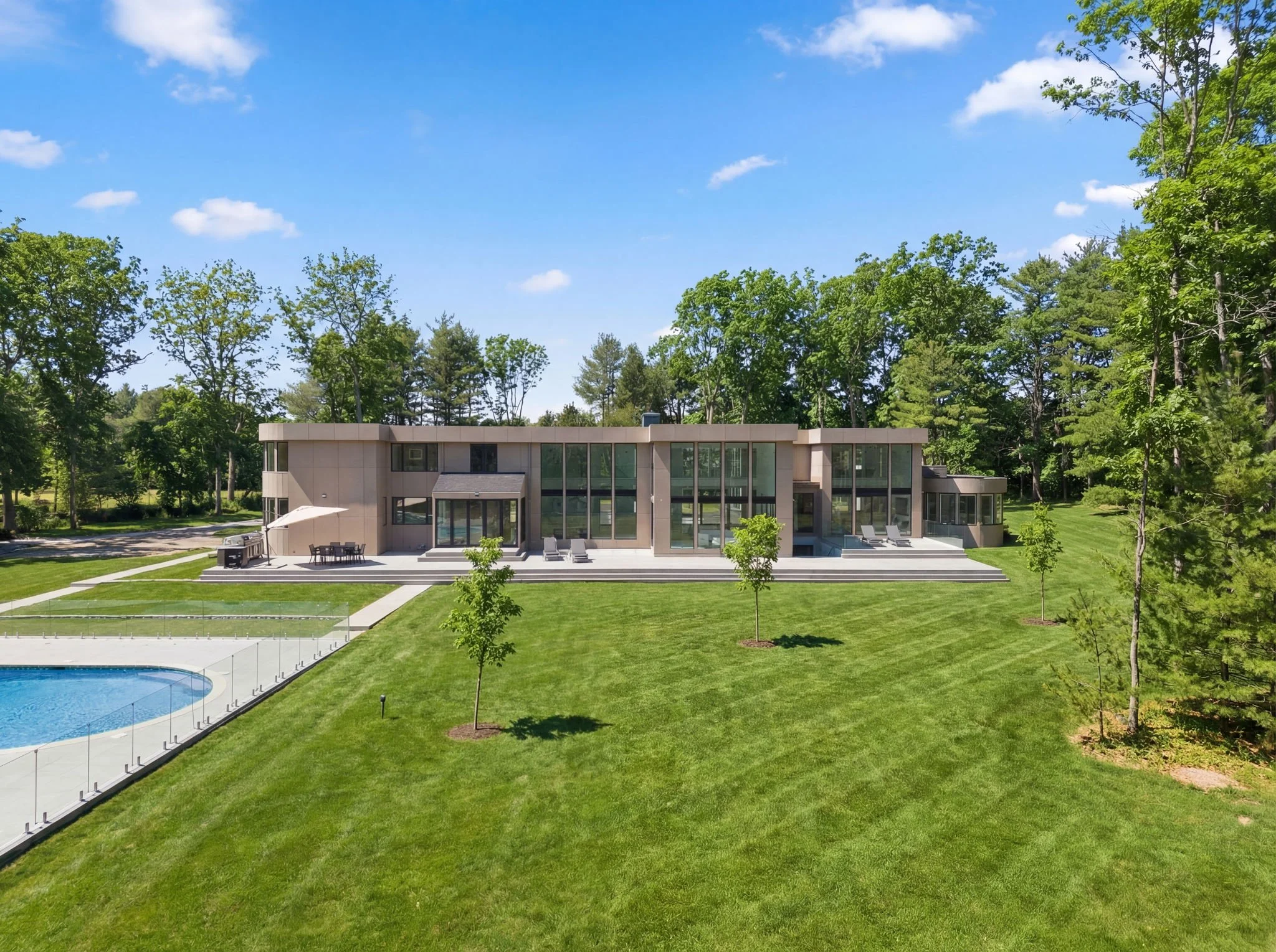 Modern house with large glass windows, surrounded by green lawn and trees, with a swimming pool in the backyard.