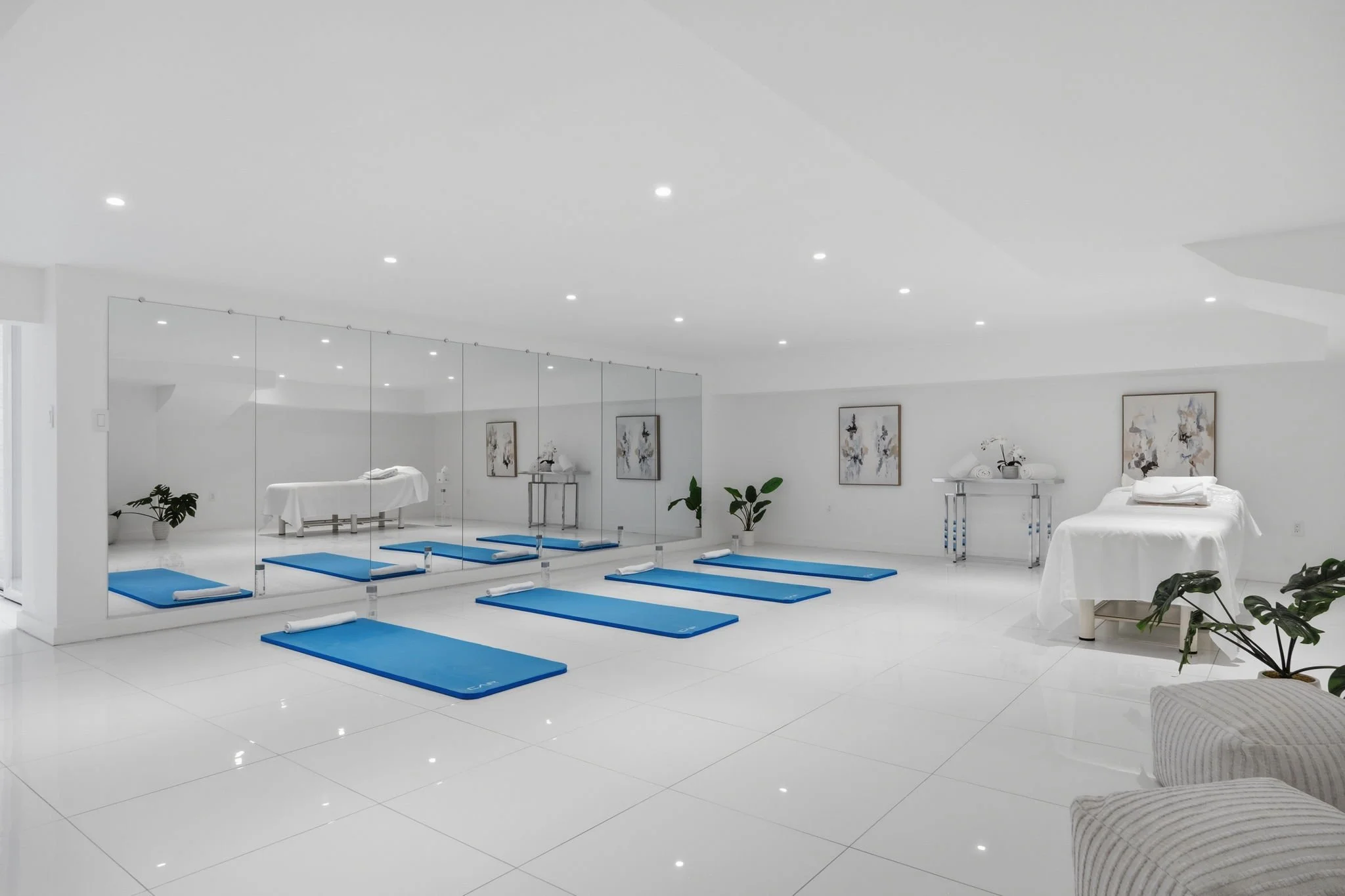 A spacious, minimalist yoga or fitness studio with white walls, ceiling, and floors. Six blue yoga mats are laid out in a row, each with a white rolled towel on top. Large mirrors cover one wall, reflecting the mats and decor. The room is decorated w