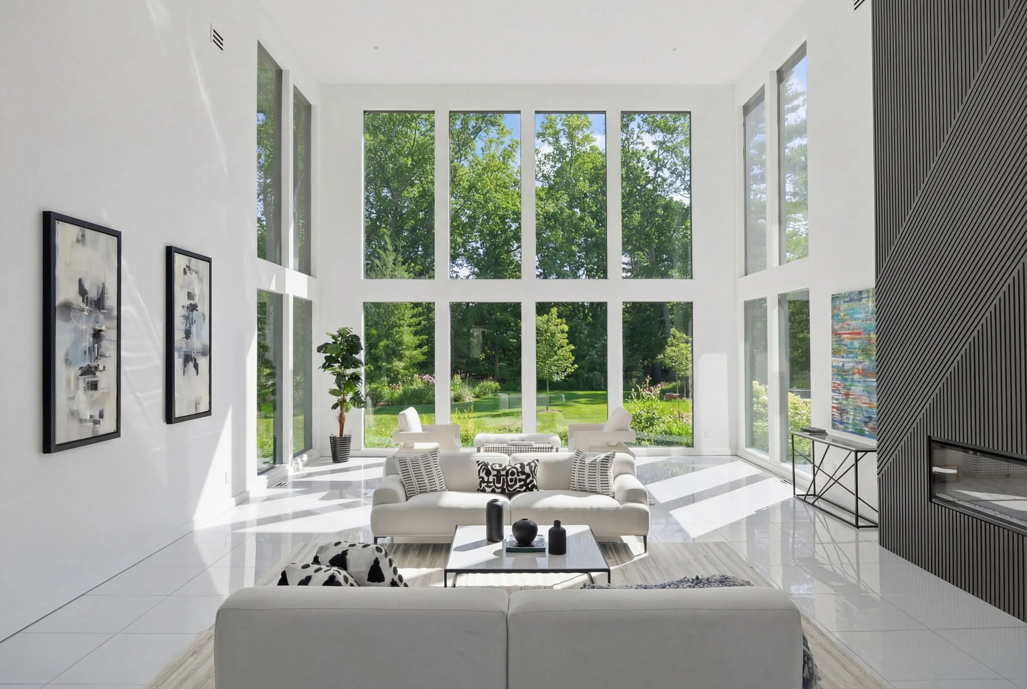 Bright, modern living room with large floor-to-ceiling windows revealing a lush green outdoor scene, white sofas, black and white decorative pillows, a black and white cow-print blanket, a glass coffee table with black and white vases, abstract art o