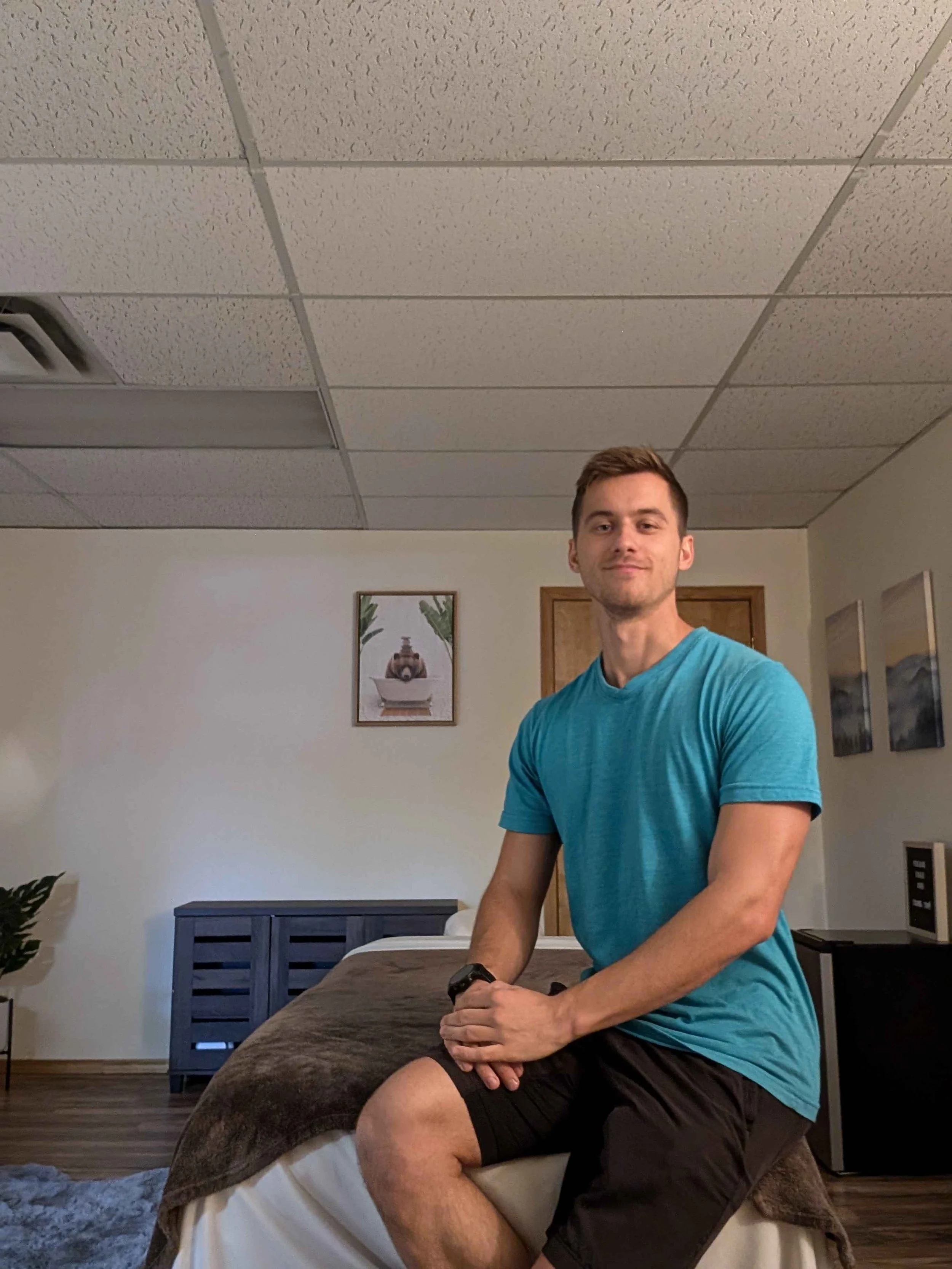 An athletically built man smiles at a camera, sitting atop a fur-covered massage table. In the background, there's a picture of a bear in a bathtub, and the cameraman (not pictured) is straining not to erupt into hysterics.