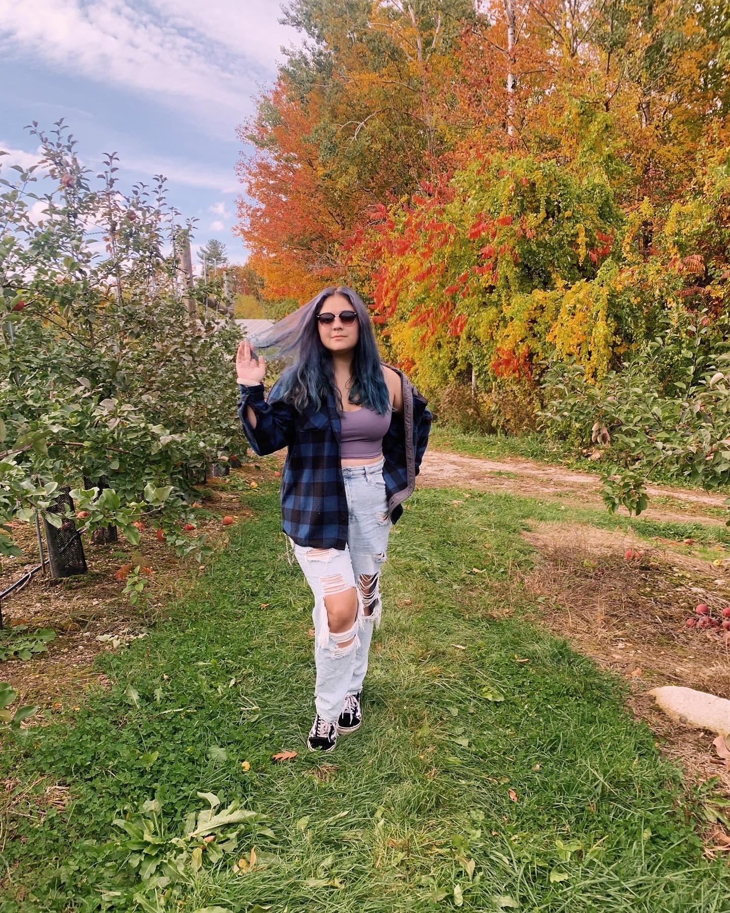 A woman with blue hair and sunglasses standing on a grassy path in an orchard with fall foliage, wearing a black and blue plaid shirt, a purple crop top, distressed jeans, and sneakers.