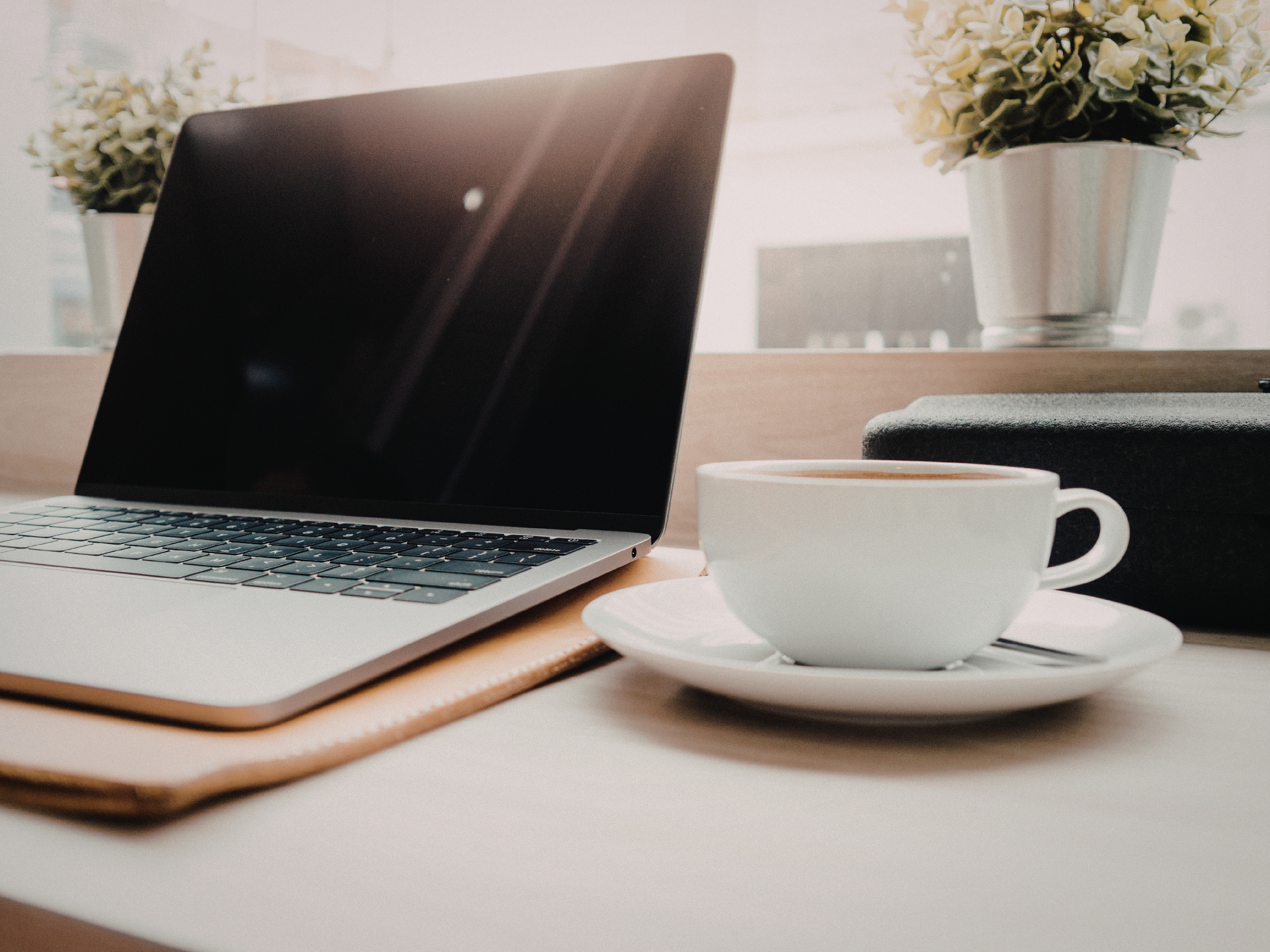 Close-up of a laptop on a desk with a coffee cup and a notebook, with potted plants in the background.