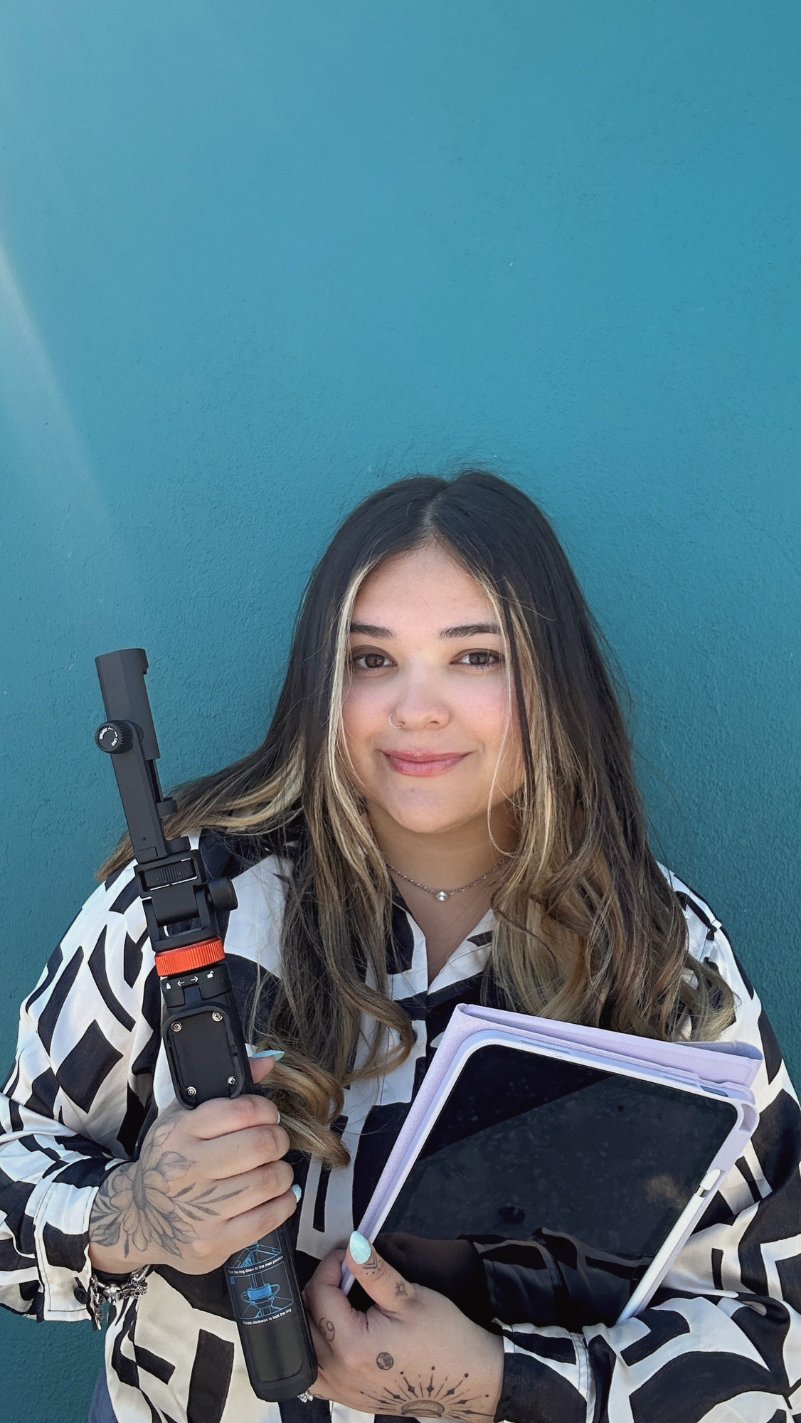 A young woman with long wavy hair, holding a film camera and a notebook, standing against a blue wall.