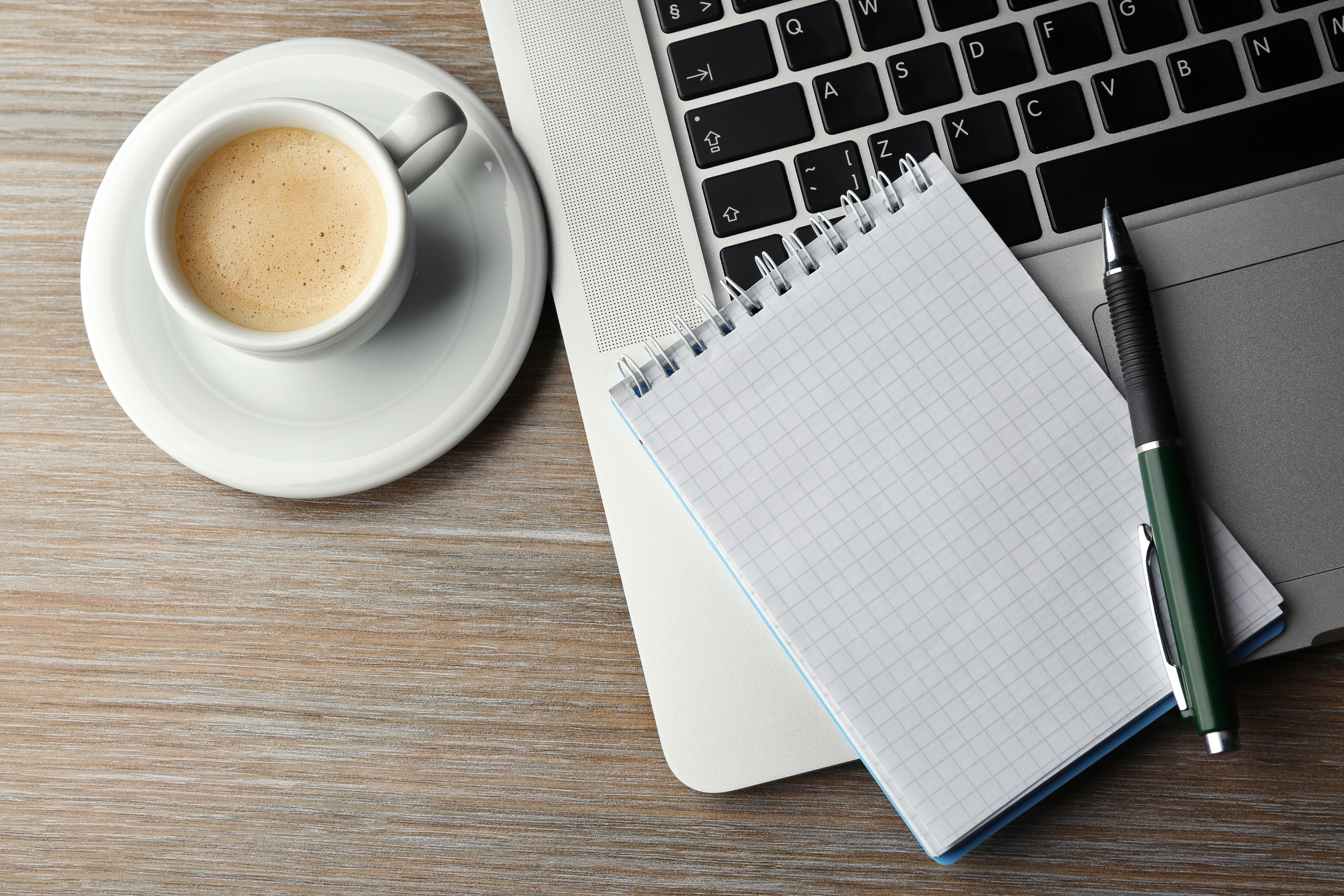 A cup of coffee on a saucer, a small spiral notepad, a pen, and a laptop on a wooden desk.