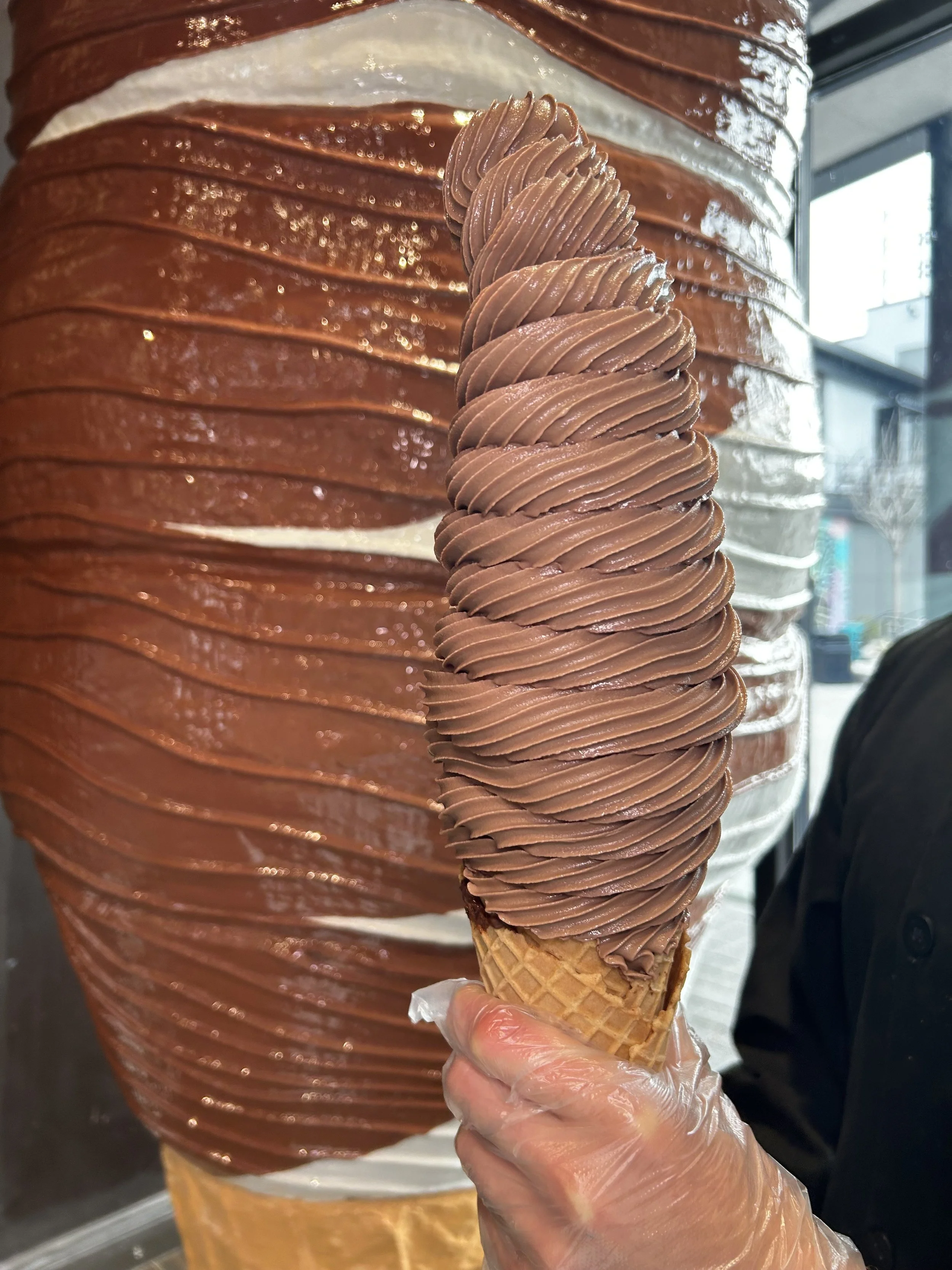 Person holding a vanilla and chocolate swirl soft serve ice cream cone with a waffle cone, in front of a large chocolate swirl ice cream mural or sculpture.
