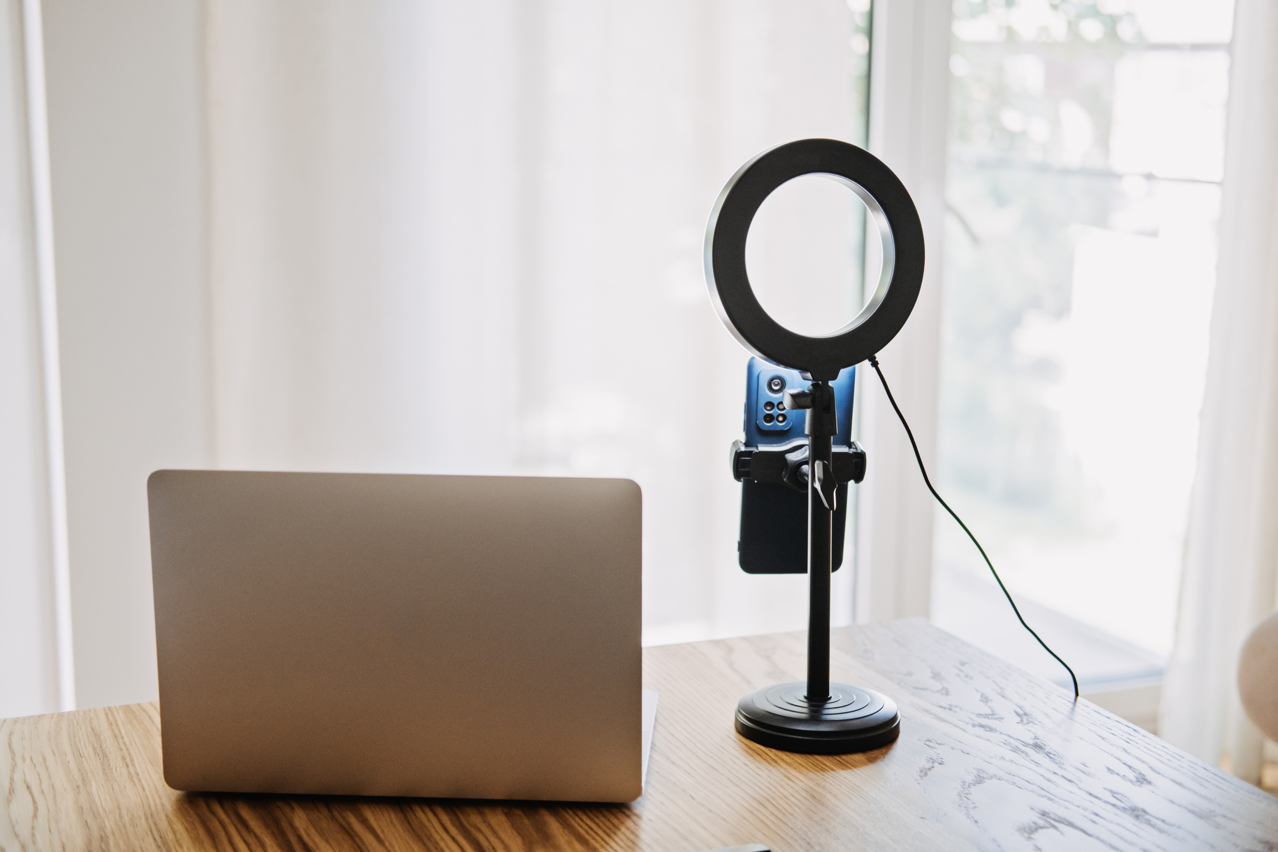 A wooden table with a silver laptop, a ring light with a phone holder, and a remote control, positioned in front of a large window with sheer curtains.