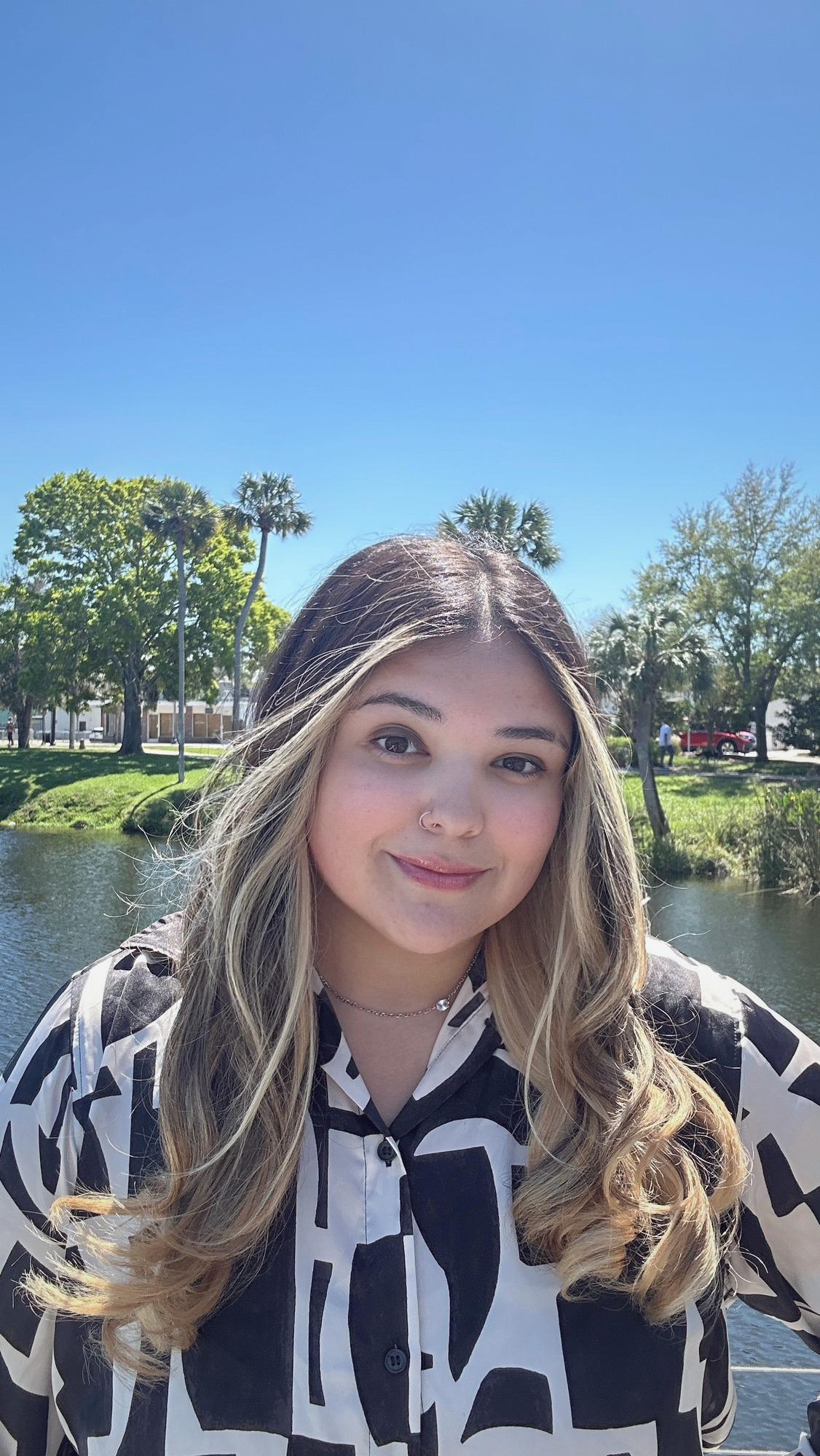 Young woman with long blonde hair, wearing a black and white patterned shirt and a nose ring, standing outdoors by a water body with trees and a blue sky in the background.