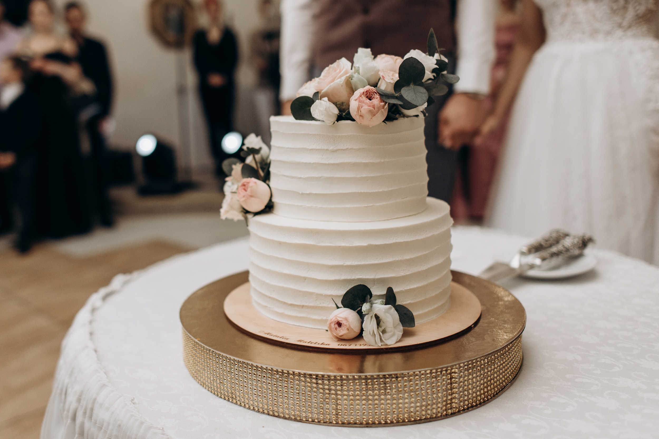 A four-tiered white wedding cake decorated with pink and white roses and greenery, displayed on a round golden cake stand at a wedding reception.