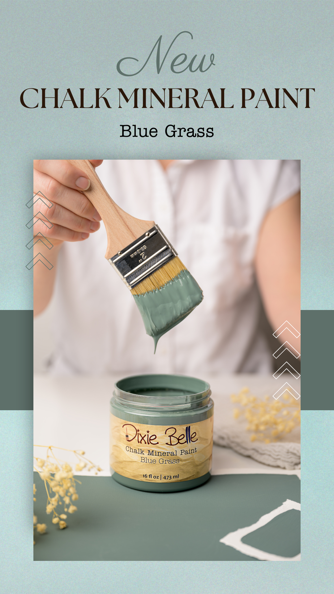 A person holding a paintbrush dipped in blue chalk mineral paint labeled "Blue Grass," with the container of the paint on a table, surrounded by flowers and a piece of white paper.