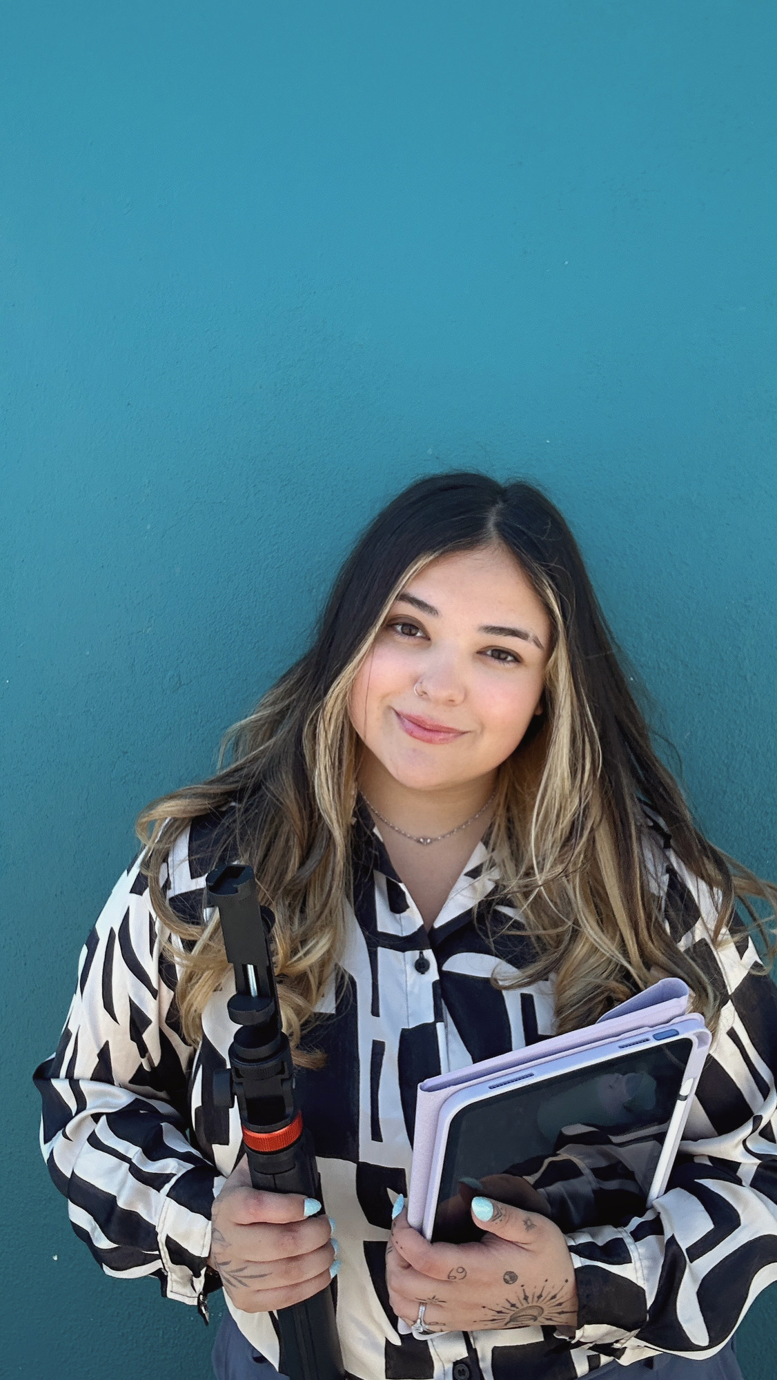 Young woman with long, wavy hair, wearing a patterned shirt, holding a smartphone and a selfie stick, standing against a teal wall.