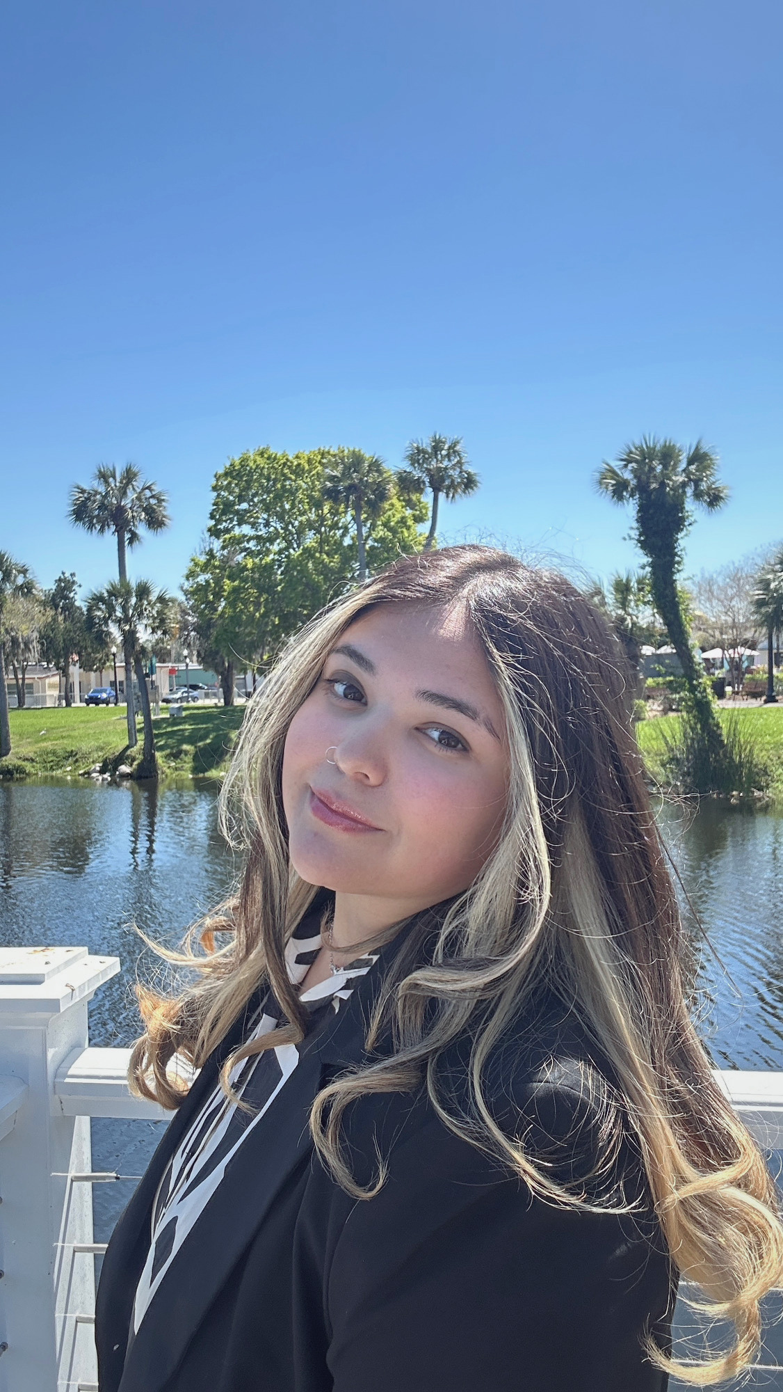 A young woman with wavy, blonde-brown hair and a nose ring, dressed in a black jacket and a striped shirt, stands near a body of water with palm trees and a blue sky in the background.