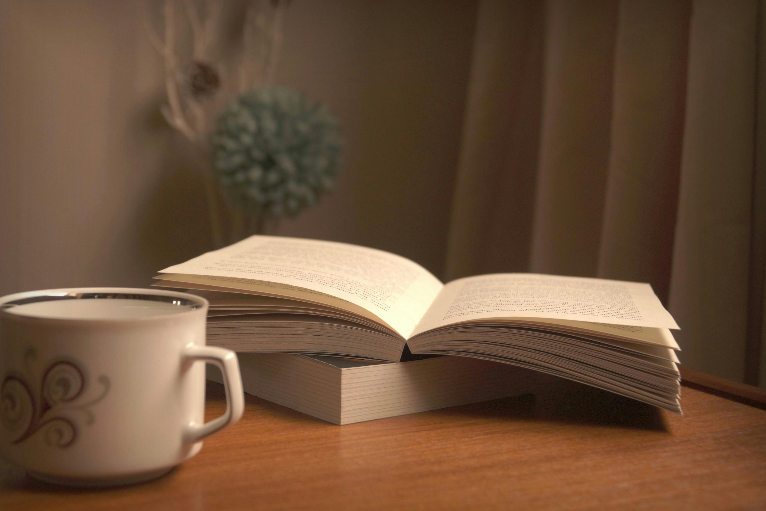 An open book with text on its pages on a wooden surface, a white coffee mug with swirl designs partially visible in the foreground, and a blurred decorative object hanging on the wall in the background.