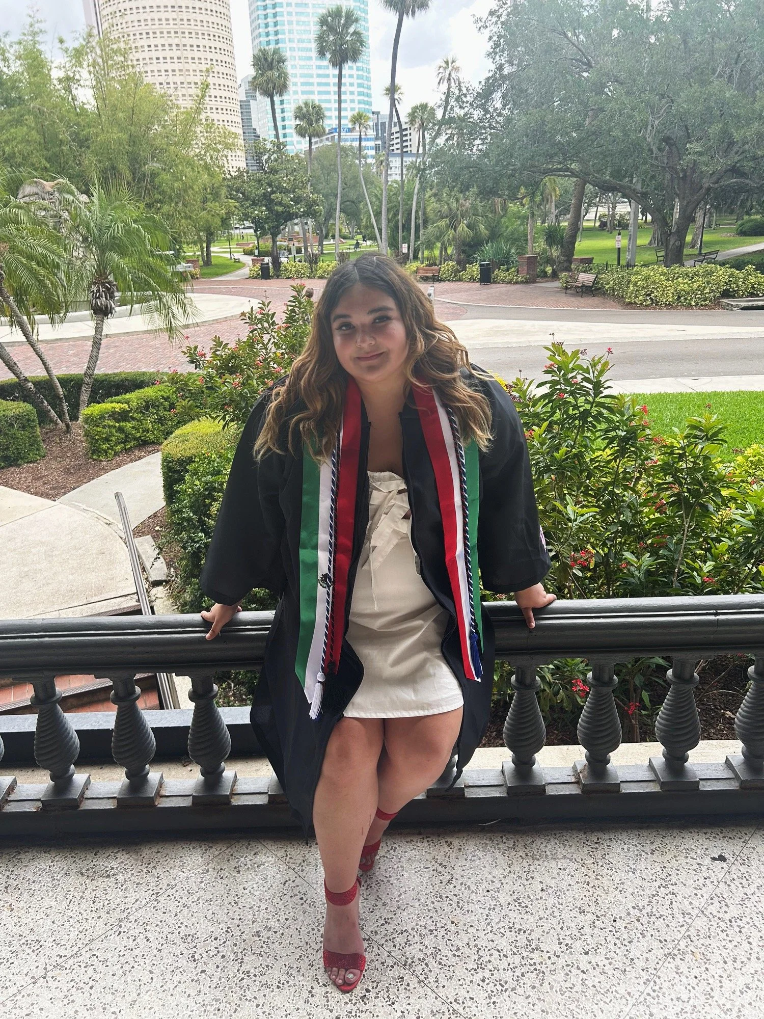Young woman in graduation gown and sash, sitting on railing outside with trees and city buildings in the background.