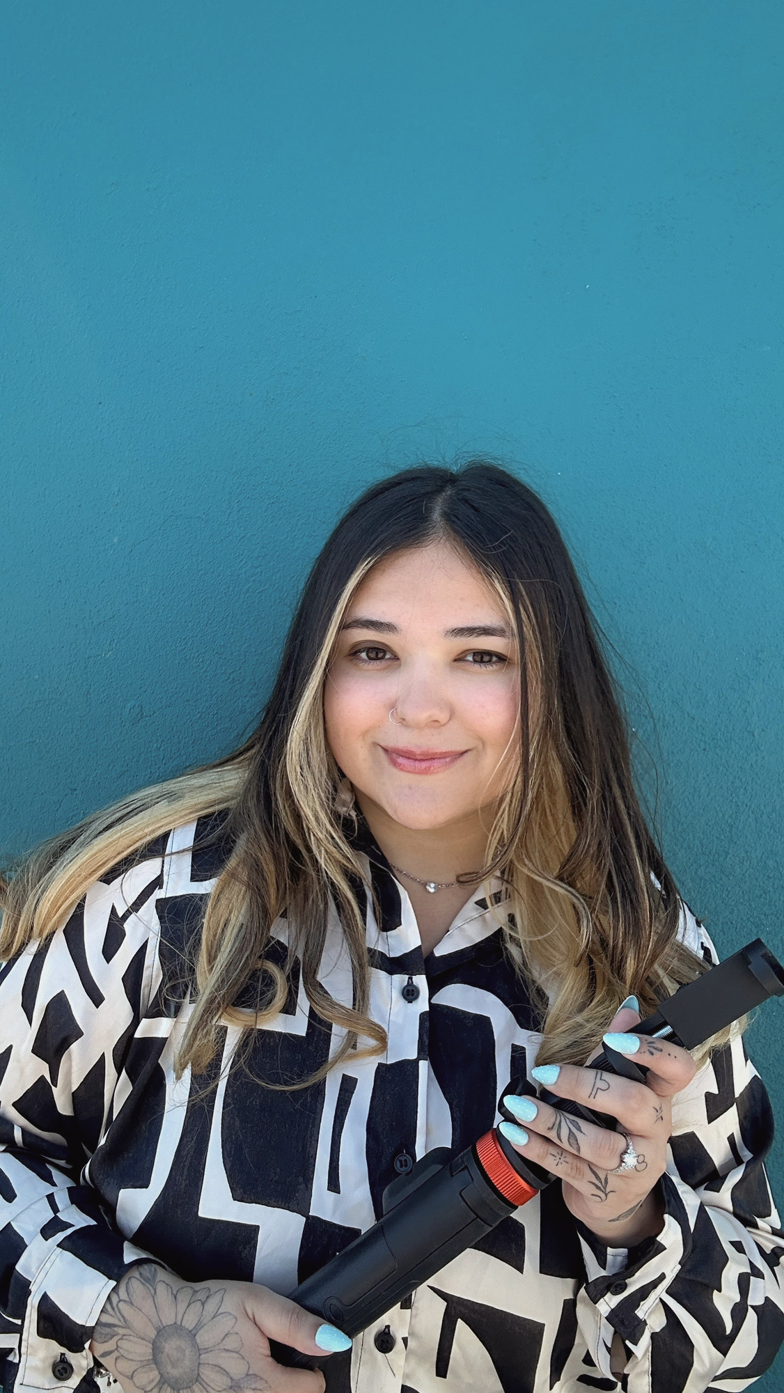 A young woman with long, wavy brown hair with blonde highlights, holding a black and red vape pen, and standing against a teal wall. She is smiling and wearing a black and white patterned shirt, with light blue nail polish, tattoos on her fingers, and a nose ring.