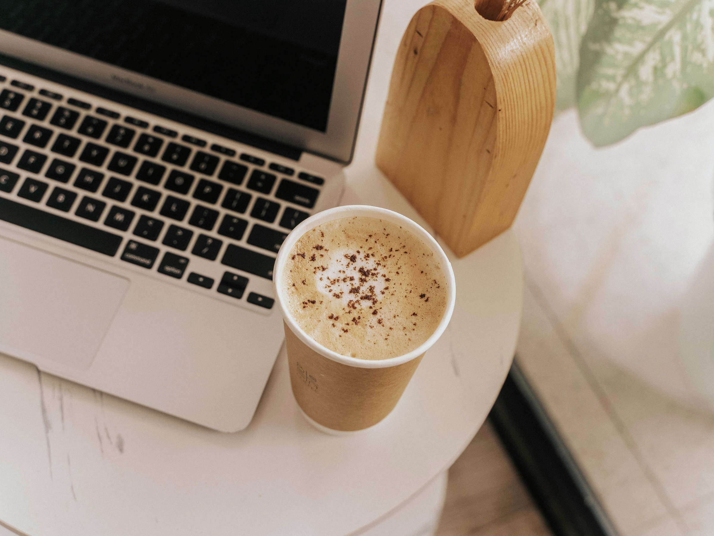 Close-up of a to-go coffee cup with frothy coffee topped with cinnamon, a laptop keyboard, a wooden tissue box, and a partially visible green leaf in the background on a white surface.