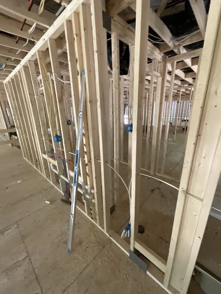 Interior of a building under construction with exposed wooden framing, electrical wiring, and construction tools leaning against the studs.