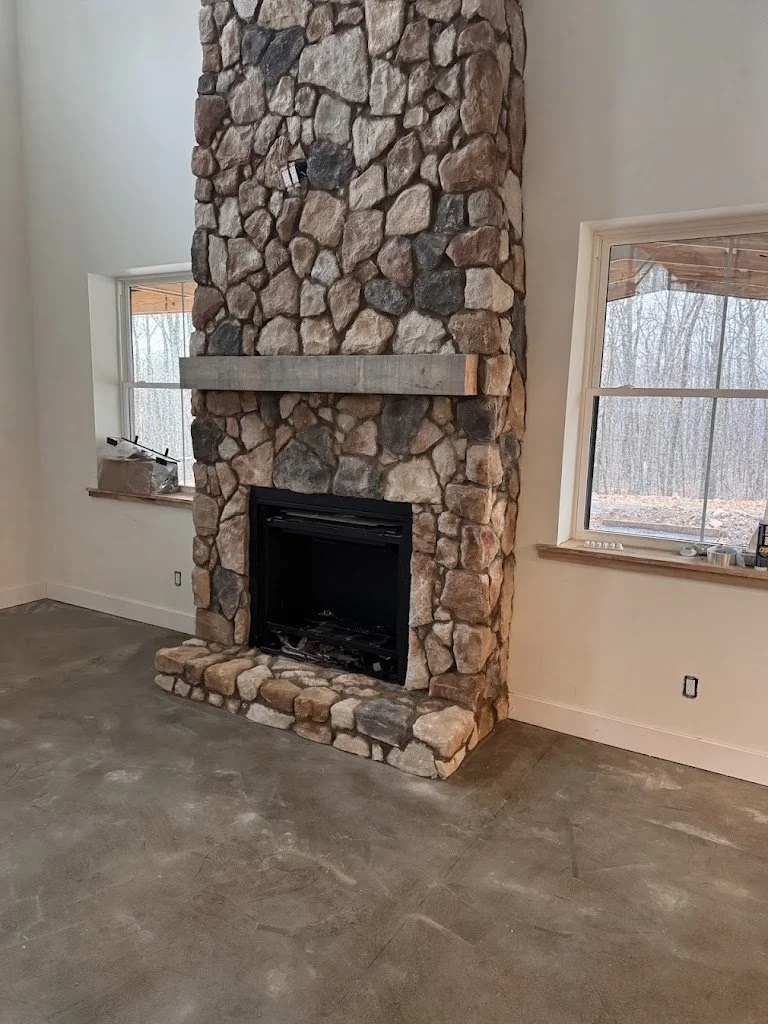 Stone fireplace with a concrete mantel in a living room with two windows and an unfinished floor.