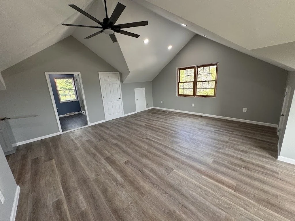 Empty living room with light gray walls, wood flooring, a large window, recessed ceiling lights, a ceiling fan, and two doorways, one leading to another room.