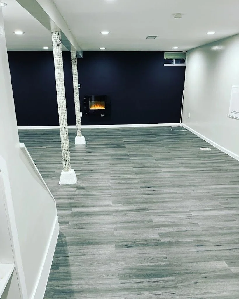 Empty basement room with wood laminate flooring, dark blue accent wall with a small electric fireplace, white walls, and ceiling with recessed lighting.