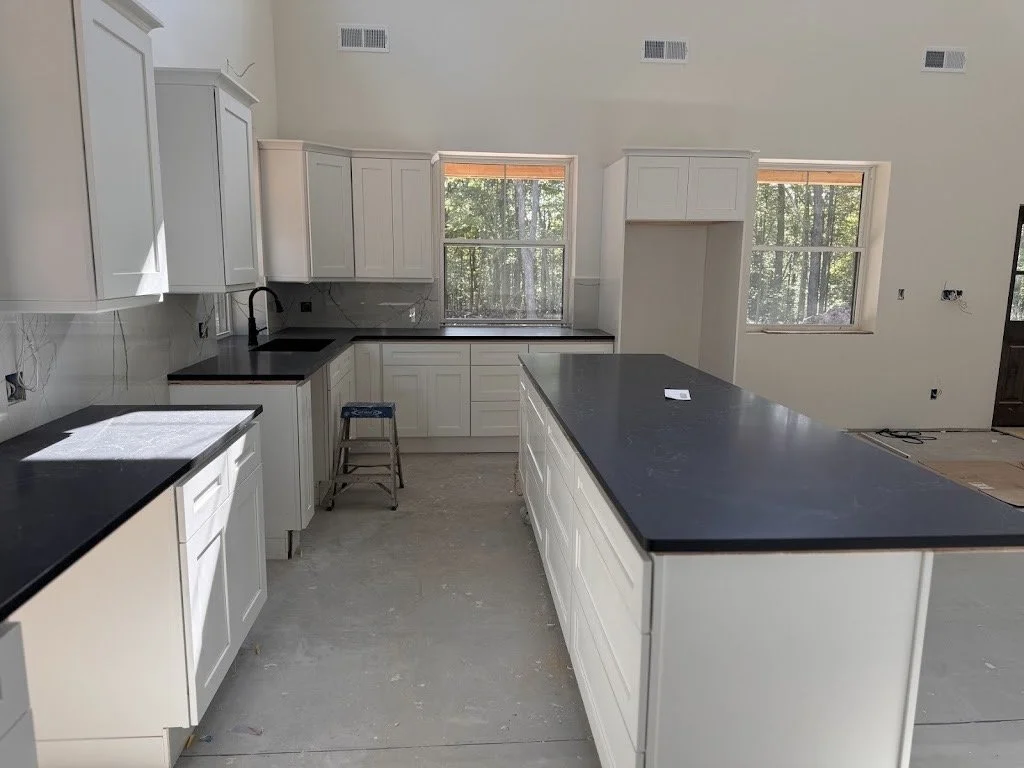 Empty kitchen with white cabinets and black countertops, a kitchen island, two windows showing trees outside, and a stool near the sink area.