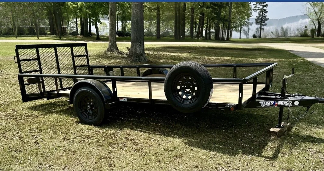 A black utility trailer with a wooden floor, parked on grass, with trees in the background.