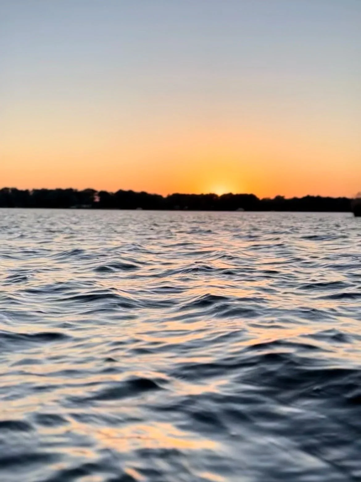 A peaceful scene of a body of water during sunset, with gentle ripples on the surface and a silhouetted treeline in the background.