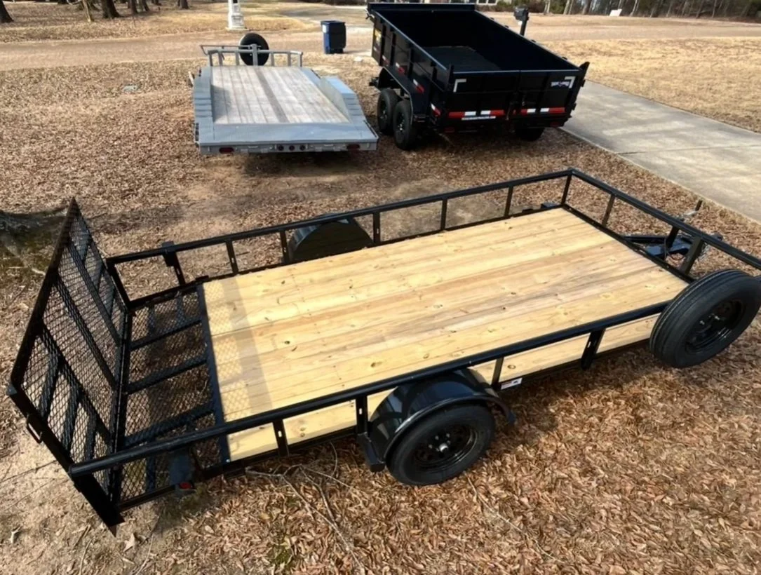 A flatbed utility trailer with a wooden deck, black metal frame, and two large black wheels parked on a leaf-covered area with other trailers in the background.