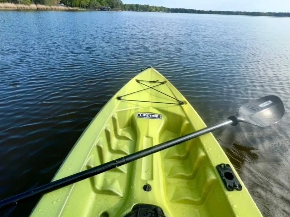 View from the front of a yellow kayak on calm water with a paddle resting across it, showing a tree-lined shoreline in the distance.