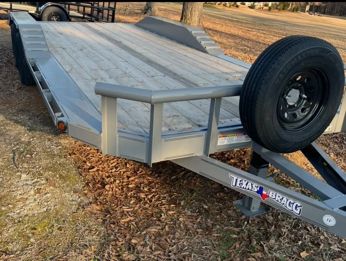 A flatbed trailer with a wooden deck, a spare tire mounted on the front right side, and a Texas Bragg logo on the frame, parked on leaf-covered ground.