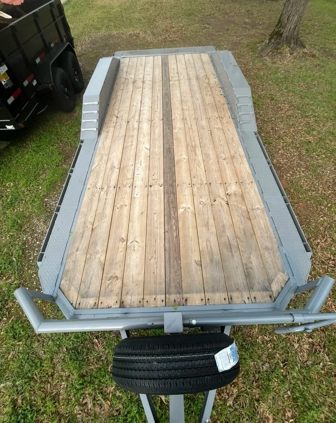 A flatbed trailer with a wooden deck parked on grass, with a black truck visible on the left side and a tree with a large trunk in the background.