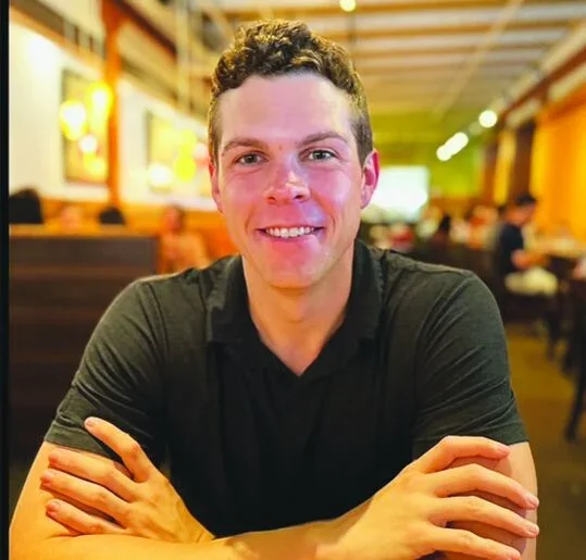 Smiling young man with short curly hair, wearing a black shirt, sitting at a table in a brightly lit restaurant.