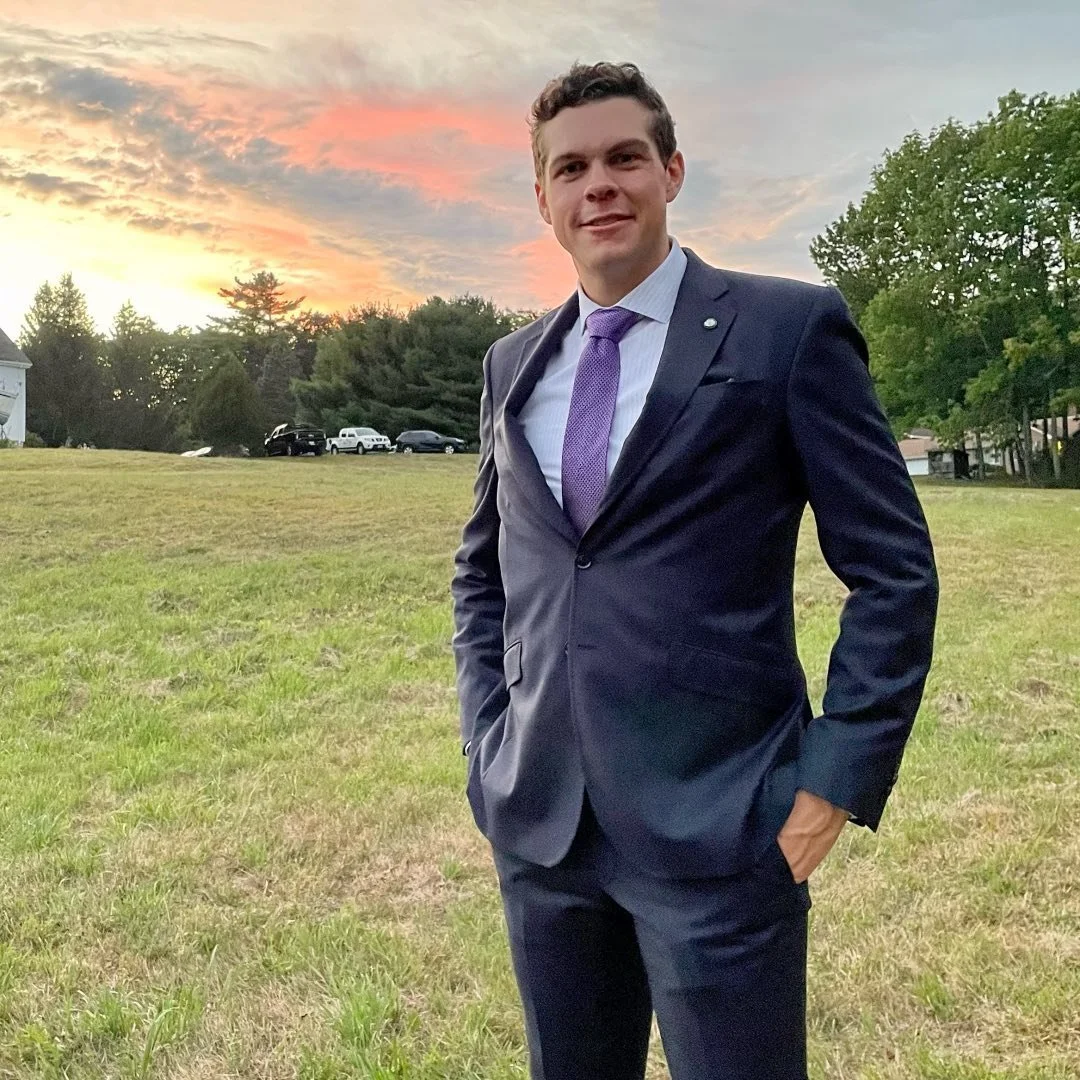 A young man in a navy blue suit with a purple tie stands outdoors on a grassy field during sunset, with trees and parked cars in the background.