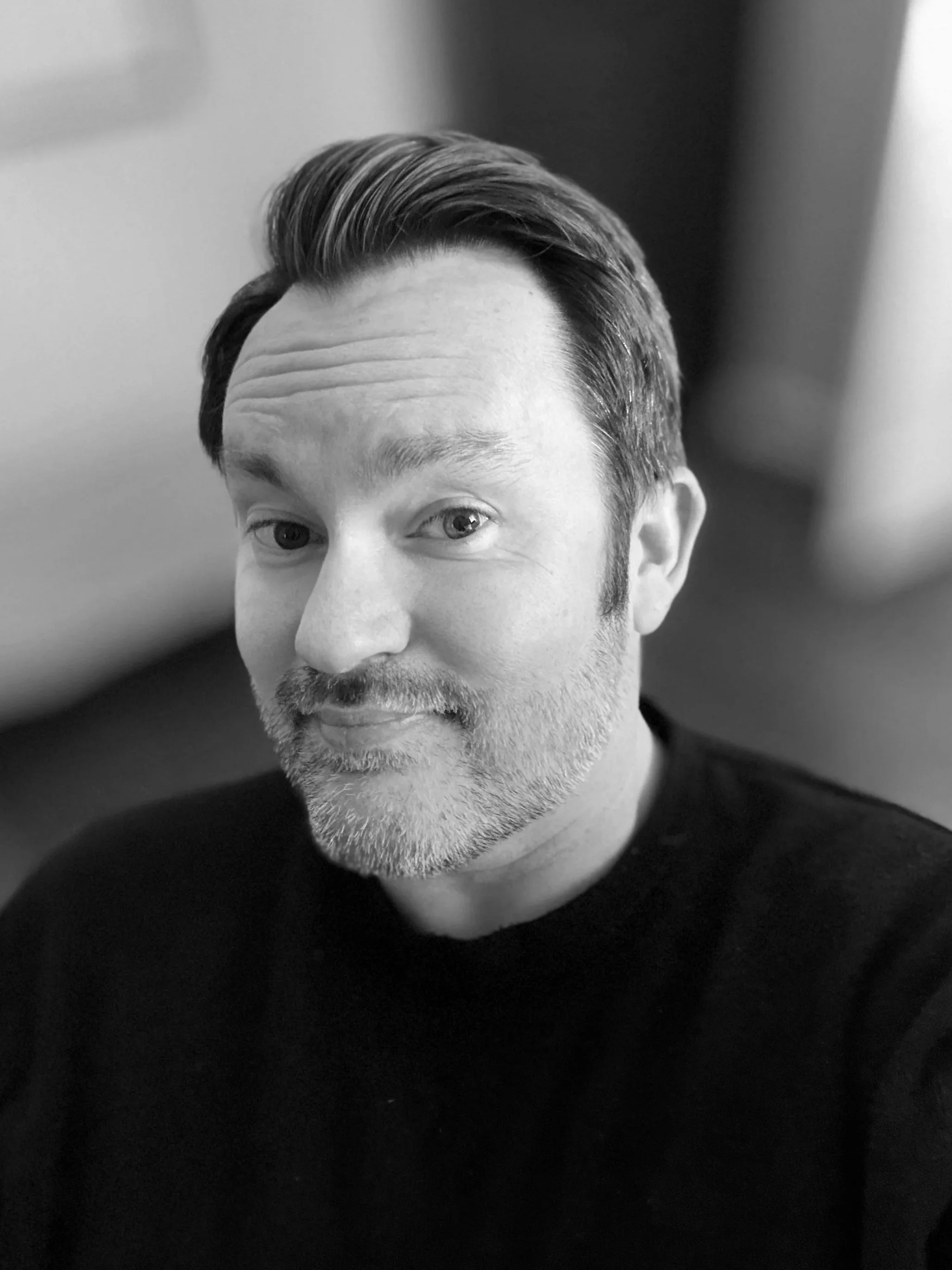 Black and white close-up of a man with styled hair, a beard, wearing a black shirt, smiling softly.