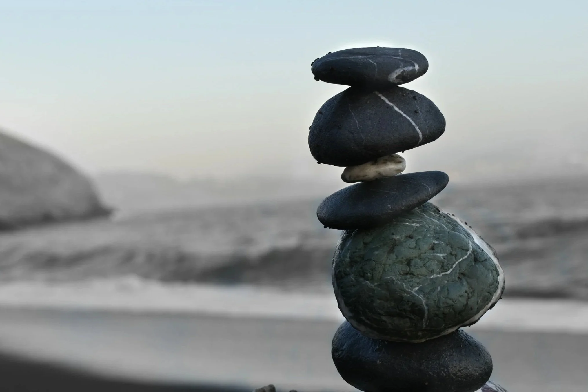 Pyramid of smooth black, gray, and white stones stacked on a beach with ocean and sky in the background.
