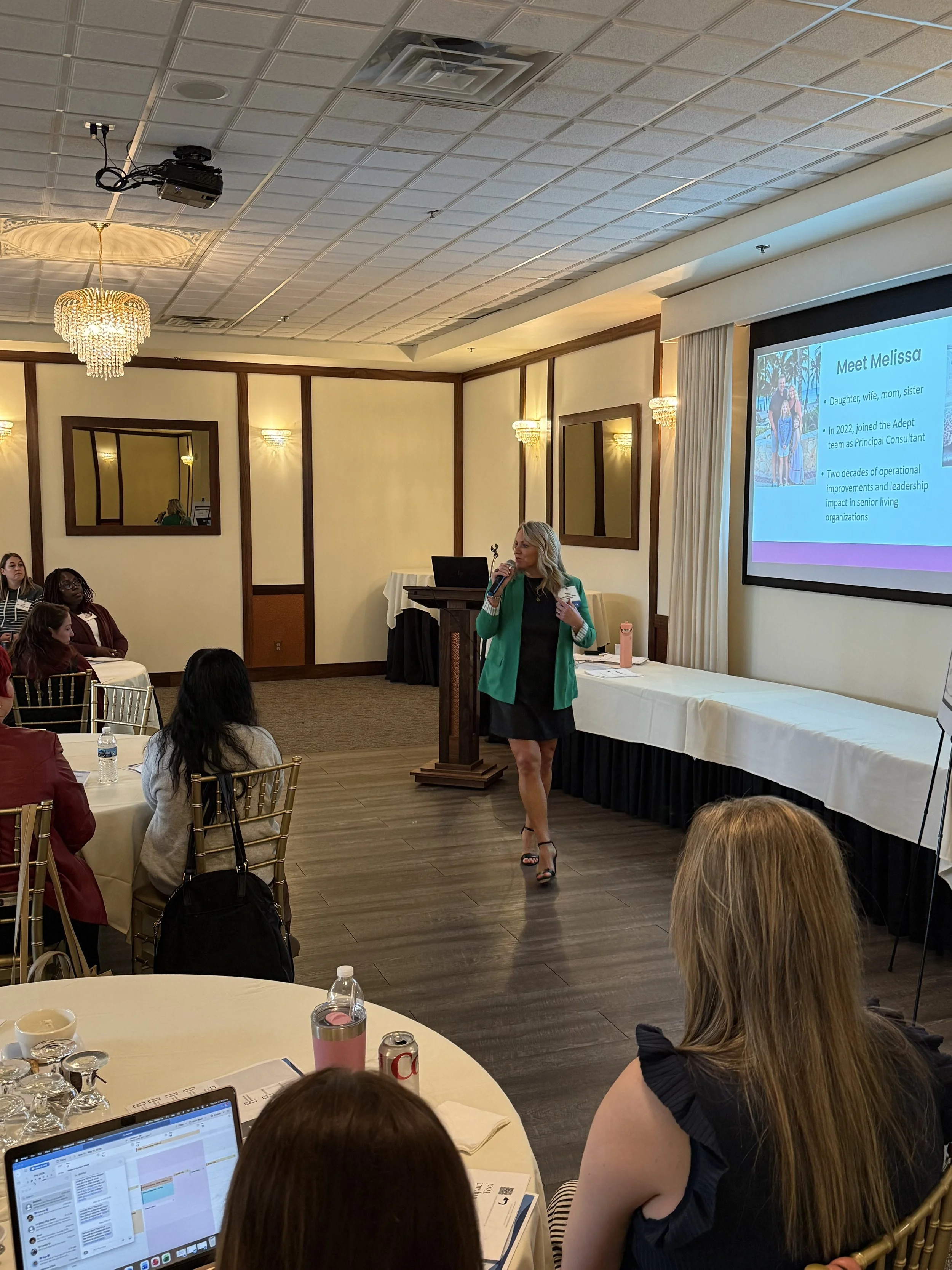 Melissa Kirchhoff, LALD, CEC, Owner and Consultant for Meridian Healthcare Strategies giving a presentation in a conference room with an audience seated at tables with large projector screen in front.