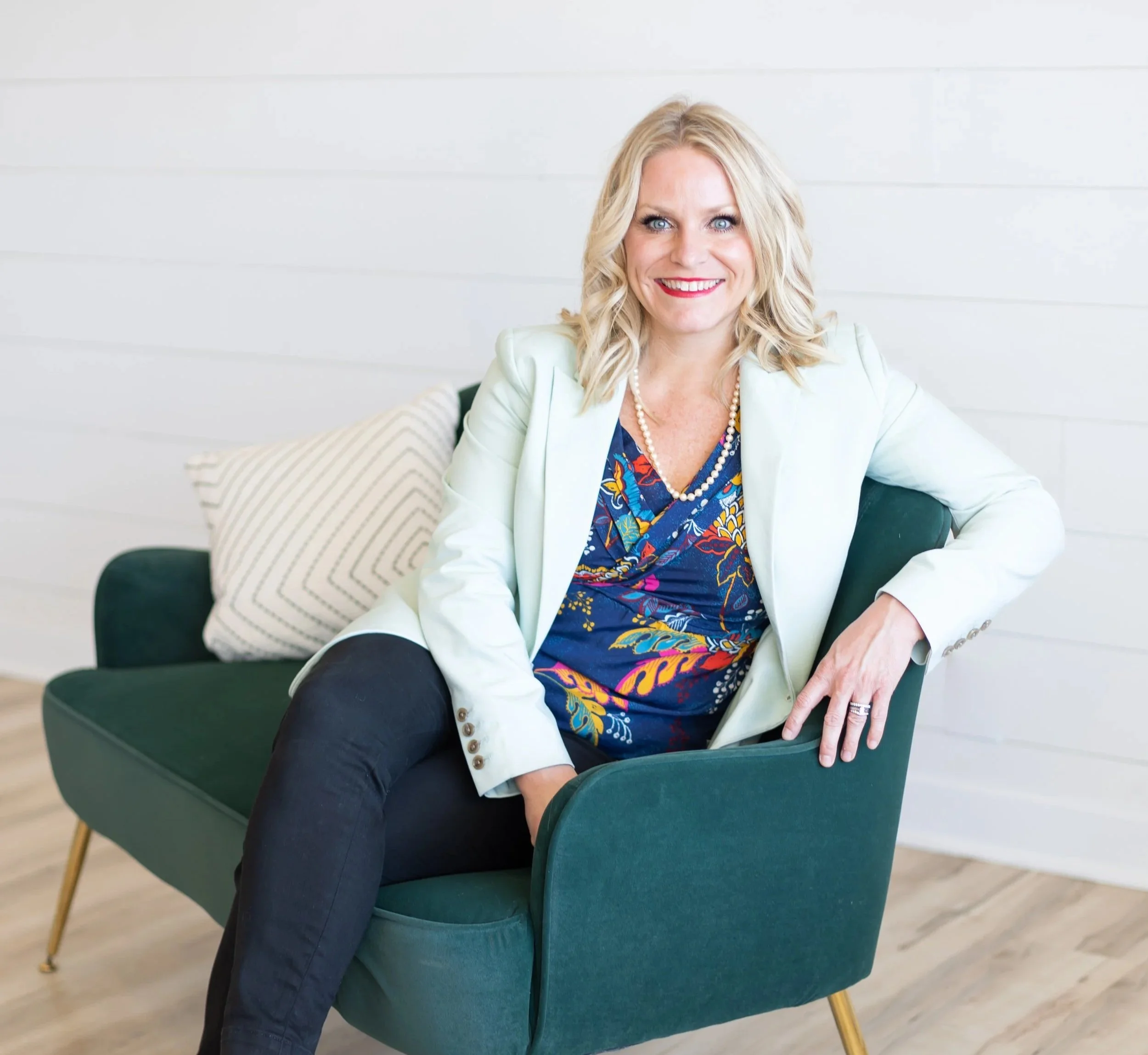 Melissa Kirchhoff, LALD, CEC, Owner and Consultant for Meridian Healthcare Strategies with blonde hair and bright blue eyes sitting on a green armchair in a bright room, smiling, wearing a white blazer, colorful dress, and pearl necklace.