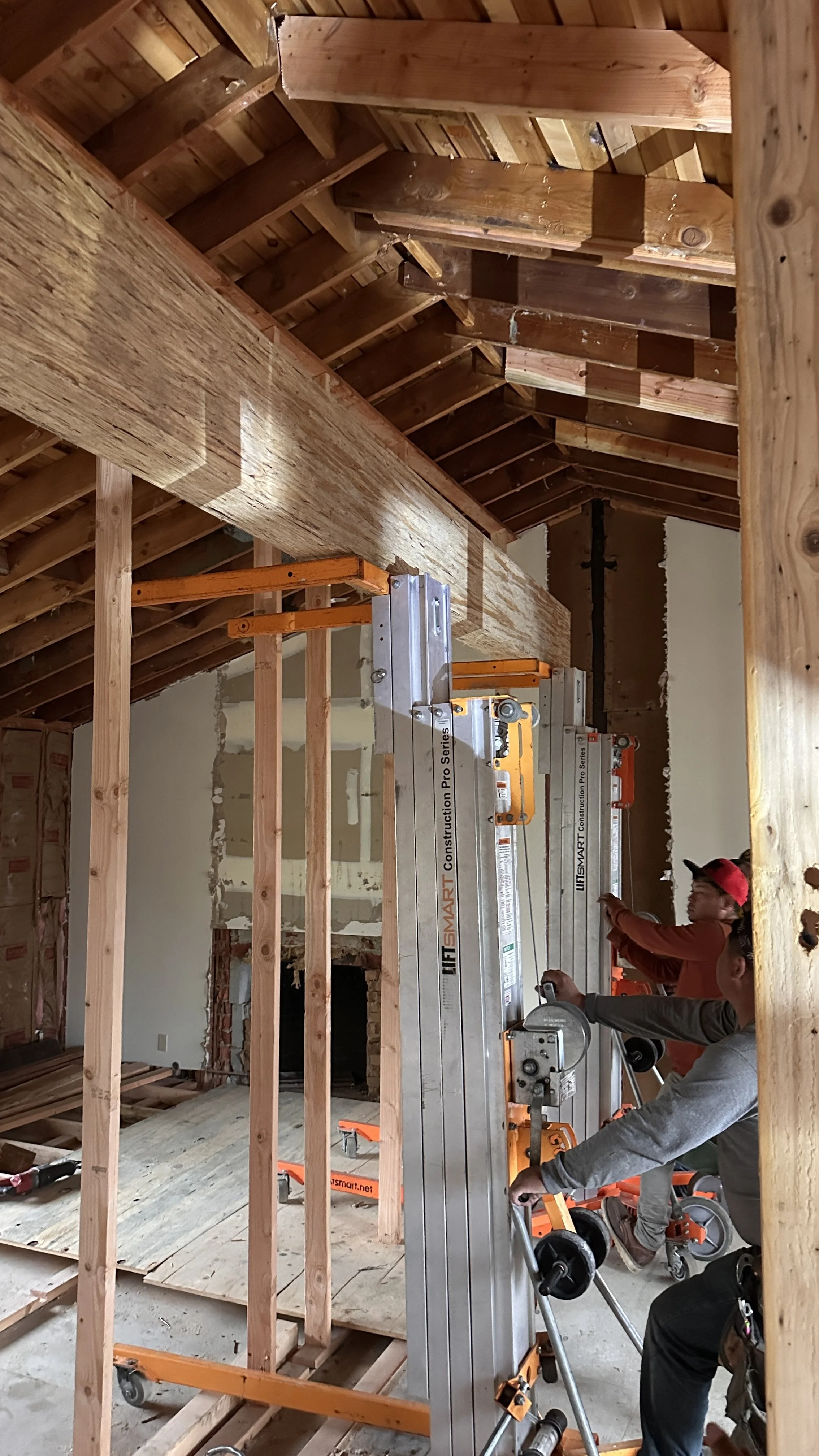 Construction workers install a large wooden beam in the framed attic of a house, using a liftsmart construction lift. The space has exposed roof rafters and unfinished interior walls.