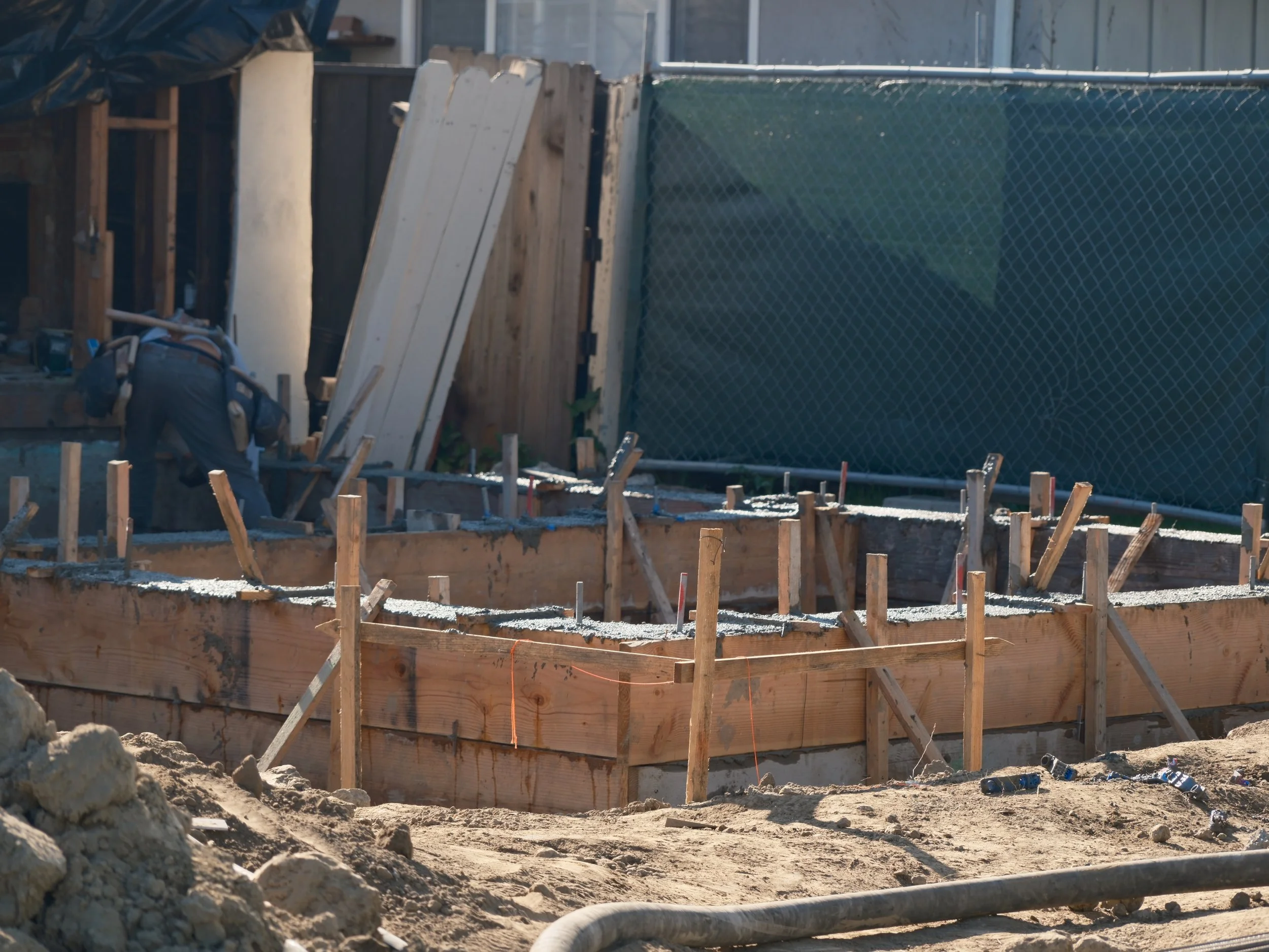 Construction site with wooden formwork for a concrete foundation, scattered tools, and a chain-link fence in the background.