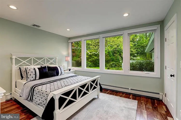 A bedroom with a white bed, black and white patterned pillows, a white linen, hardwood floors, and large windows overlooking greenery.