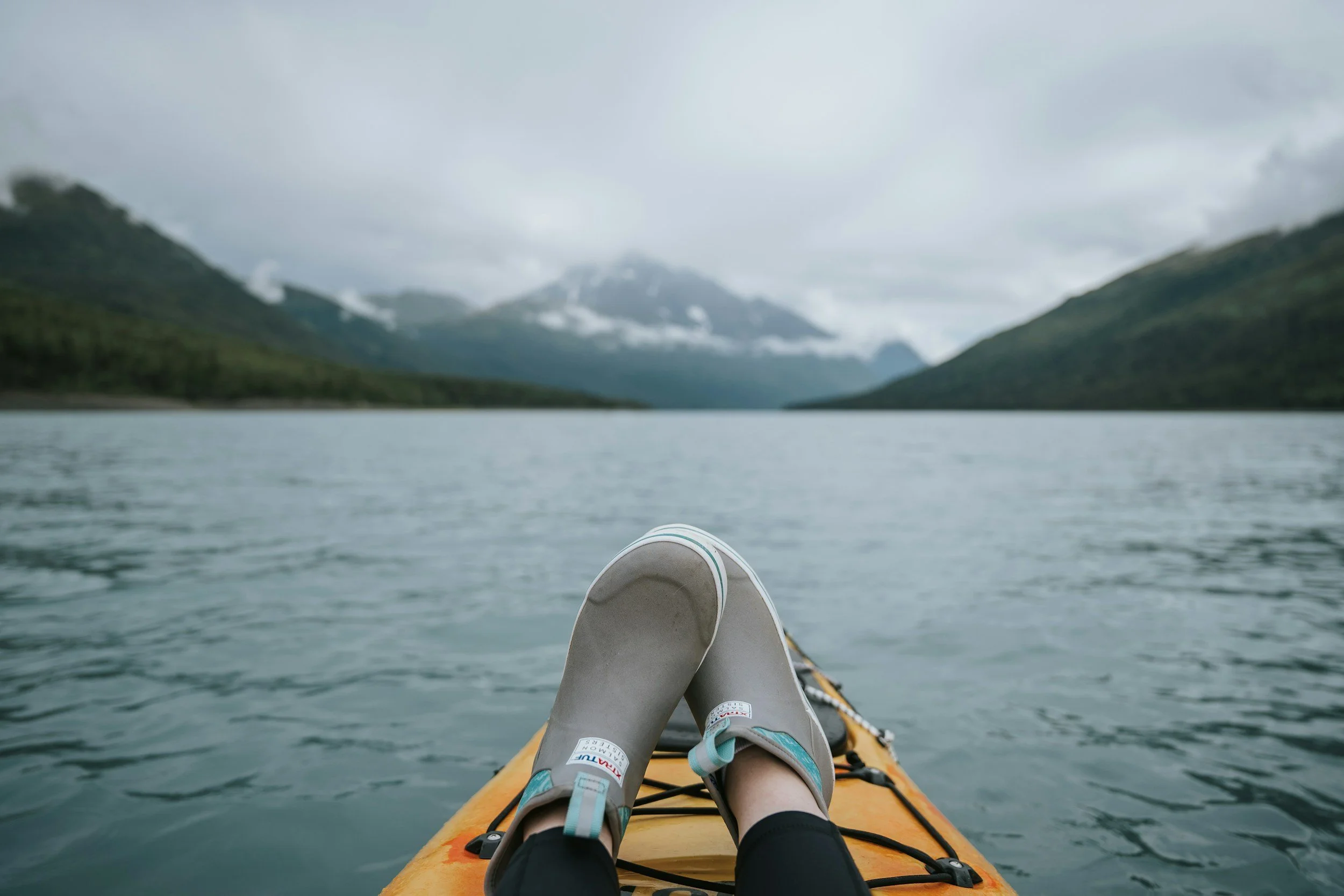 Person relaxing on a kayak on a lake with mountains and cloudy sky in the background