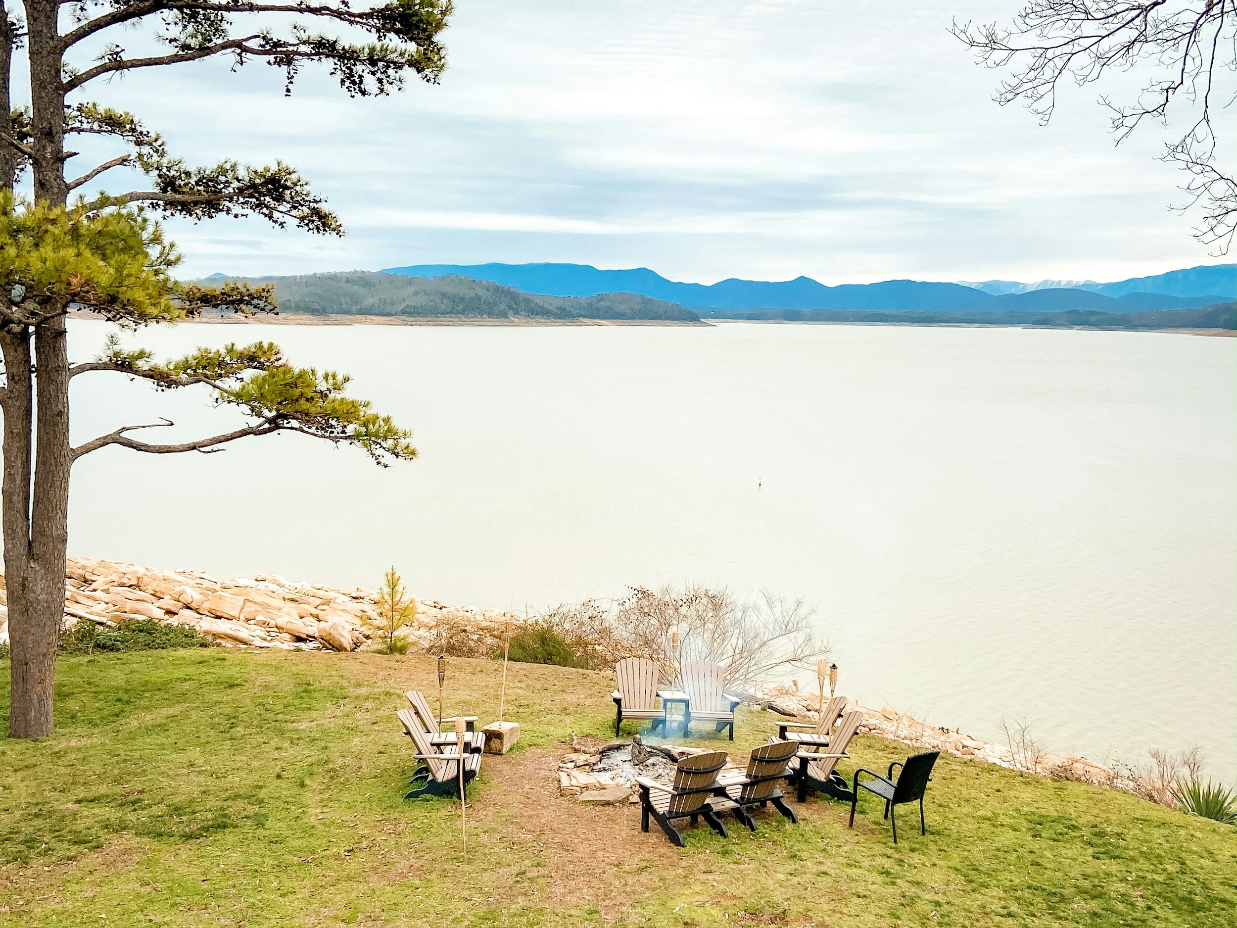 A lakeside backyard with a campfire area, lawn chairs, and Adirondack chairs arranged around a fire pit, overlooking a large body of water with hills and mountains in the distance.