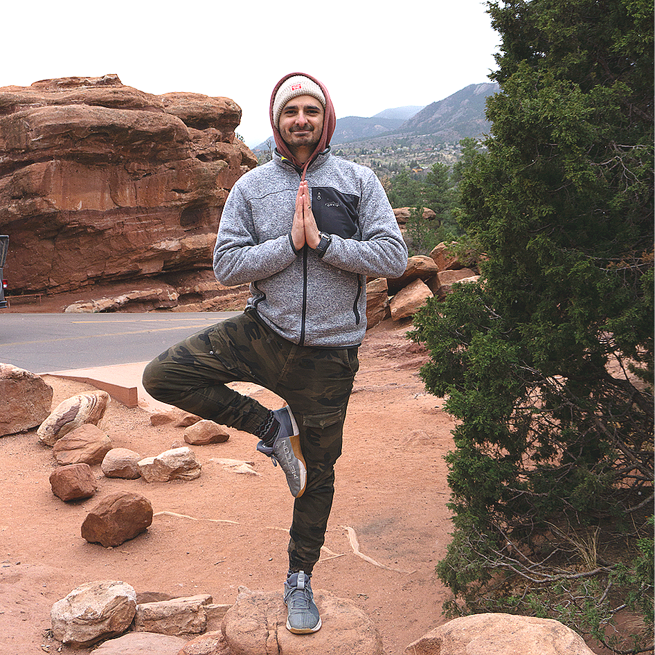 Man in gray hoodie and camouflage pants practicing yoga in a rocky outdoor landscape, balancing on one leg with hands in prayer position.
