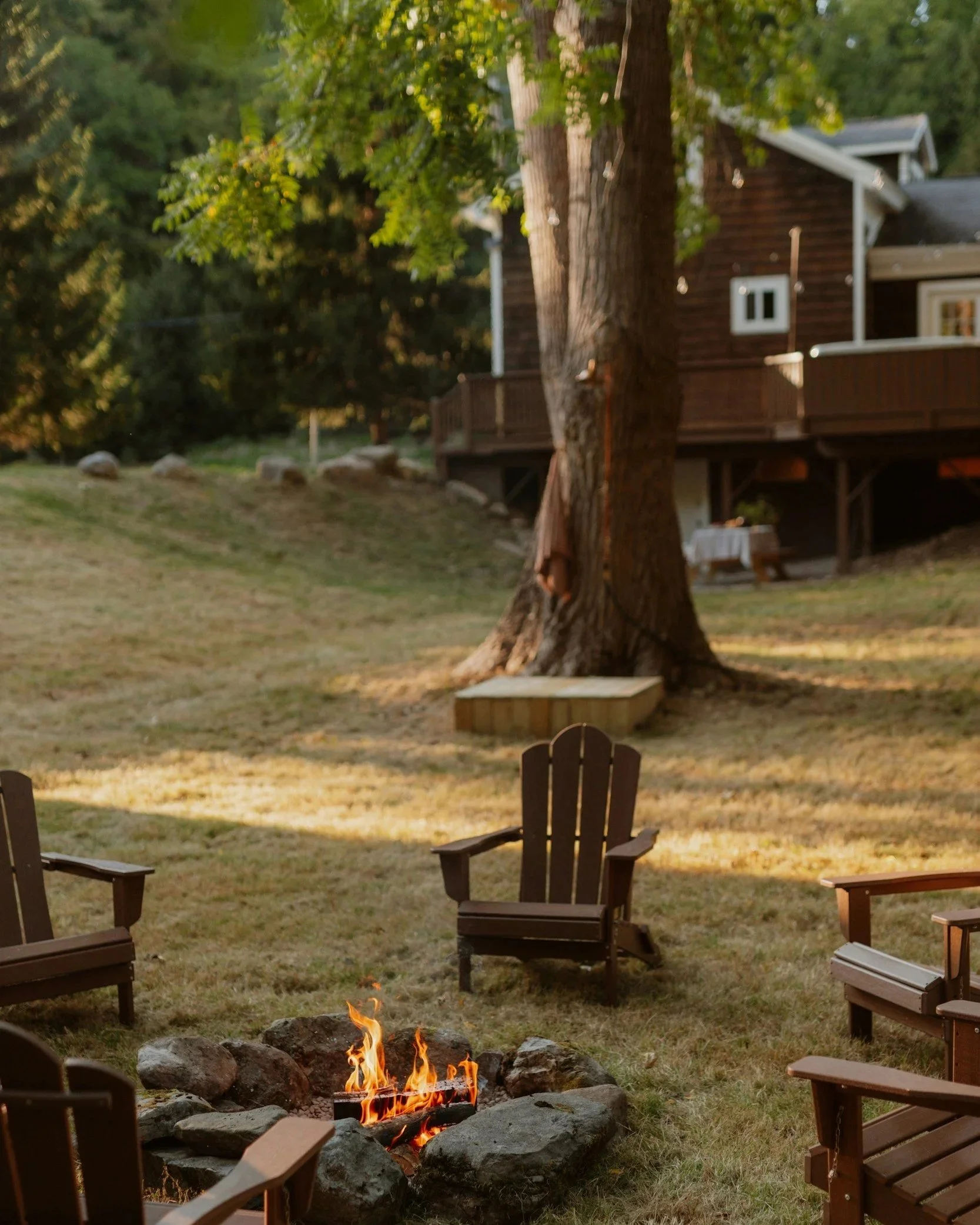 A backyard outdoor fire pit with flames surrounded by Adirondack chairs at sunset, trees, and a house in the background.