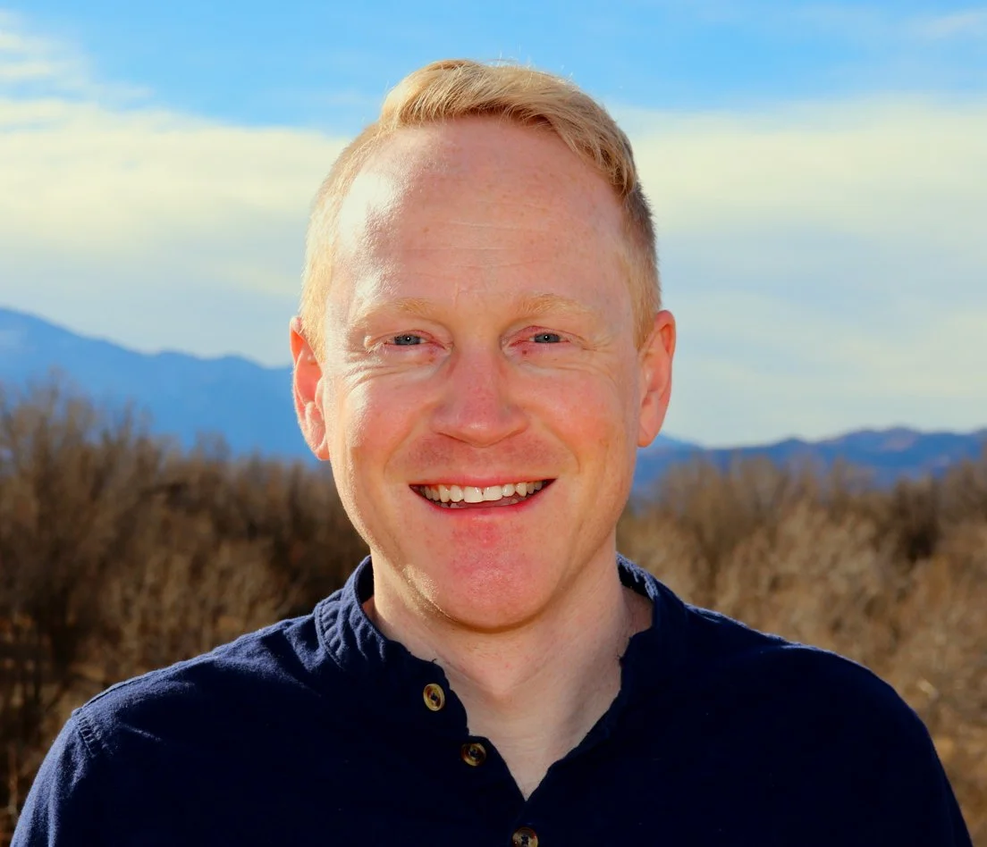 A smiling man with short blond hair, wearing a dark button-up shirt, outdoors with mountains and blue sky in the background.