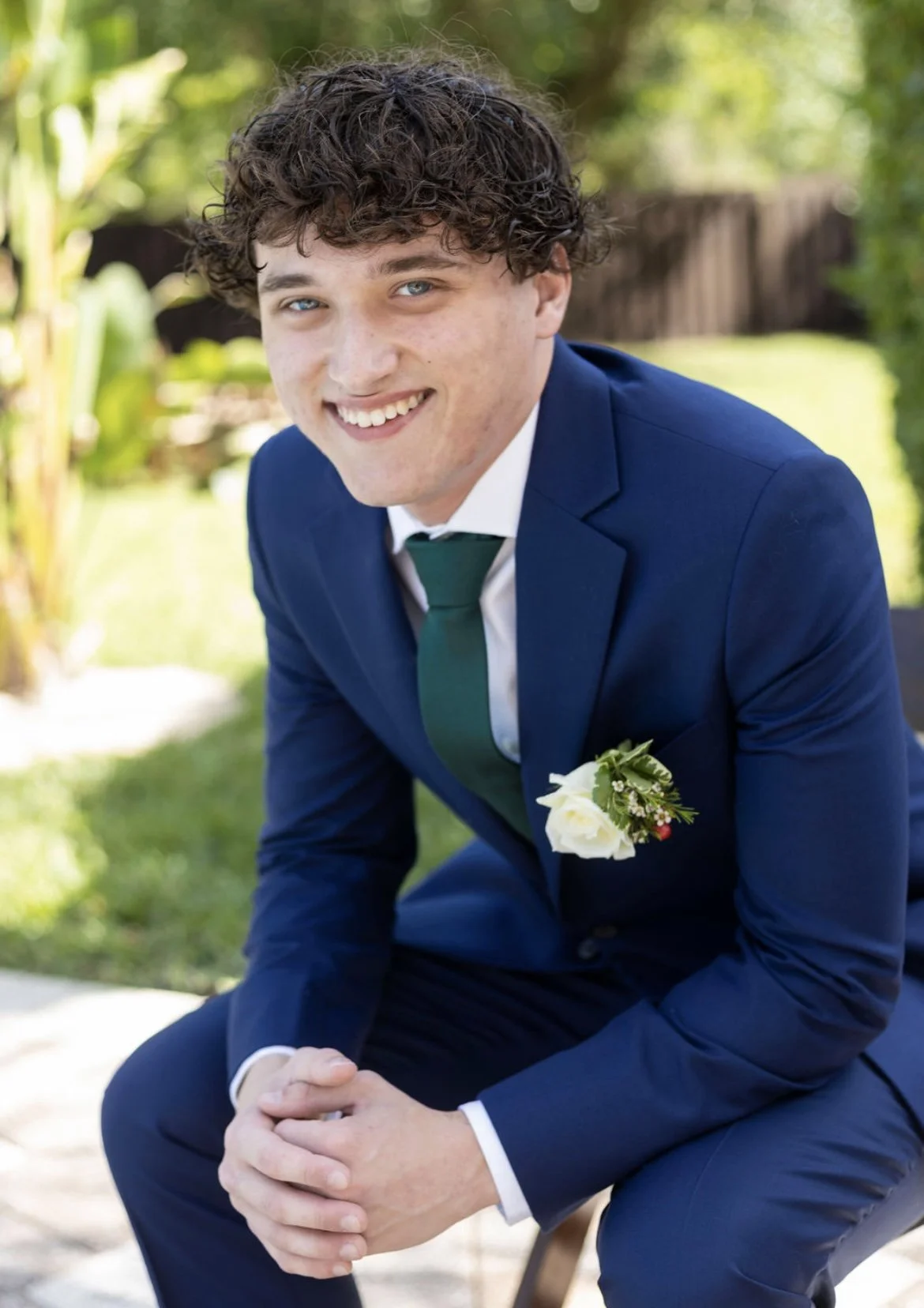 Young man with curly hair wearing a blue suit, white shirt, and green tie, smiling outdoors, with a white flower boutonniere on his lapel.
