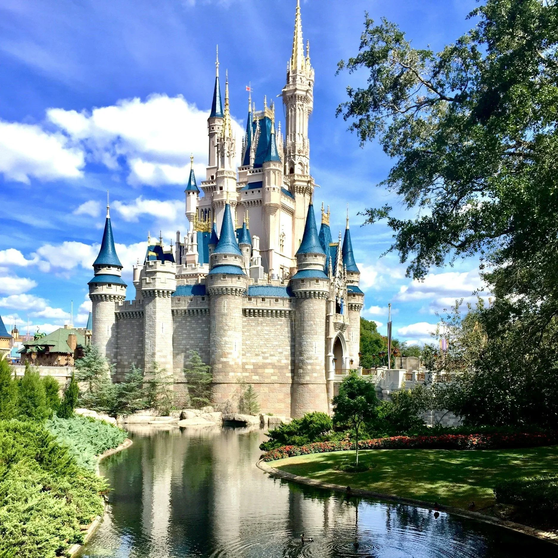 A large castle with multiple towers and blue roofs, situated by a reflective body of water, with trees and a partly cloudy sky in the background.