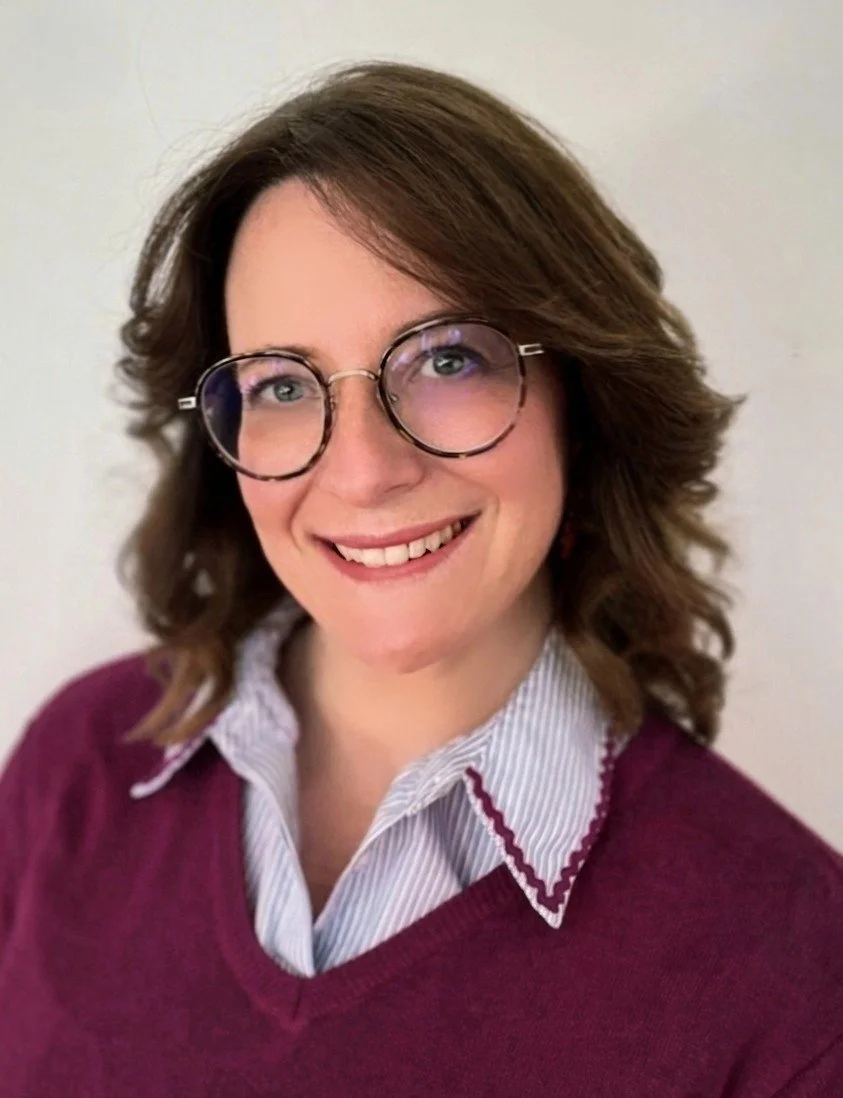 Anne Sophie Aparicio : une femme aux cheveux bruns, portant des lunettes rondes, un pull violet et une chemise rayée blanche et bleue, sourit devant un fond neutre.